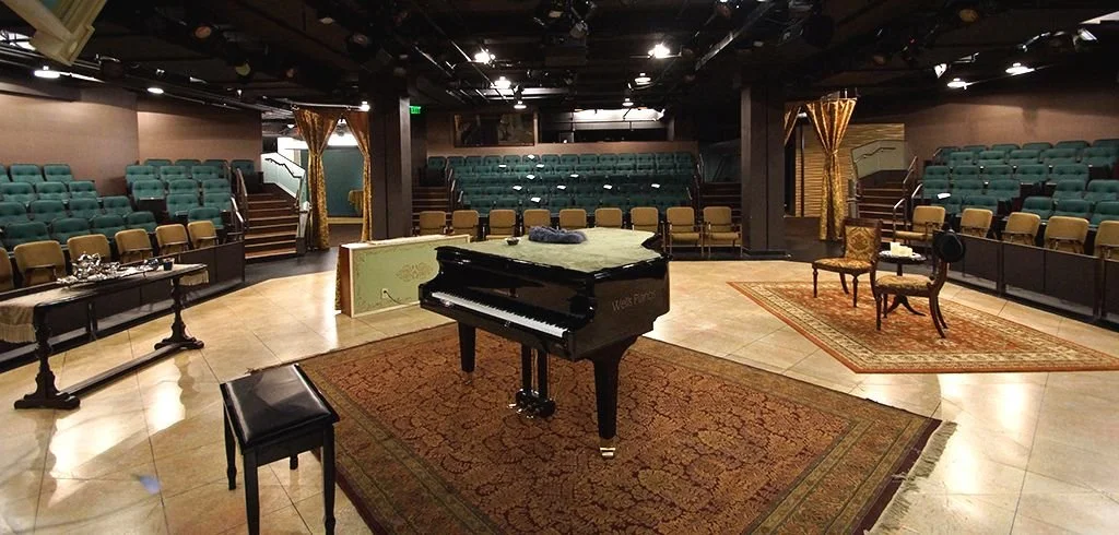 Empty theater stage with a grand piano, two chairs, and a table with silverware, surrounded by rows of green theater seats.