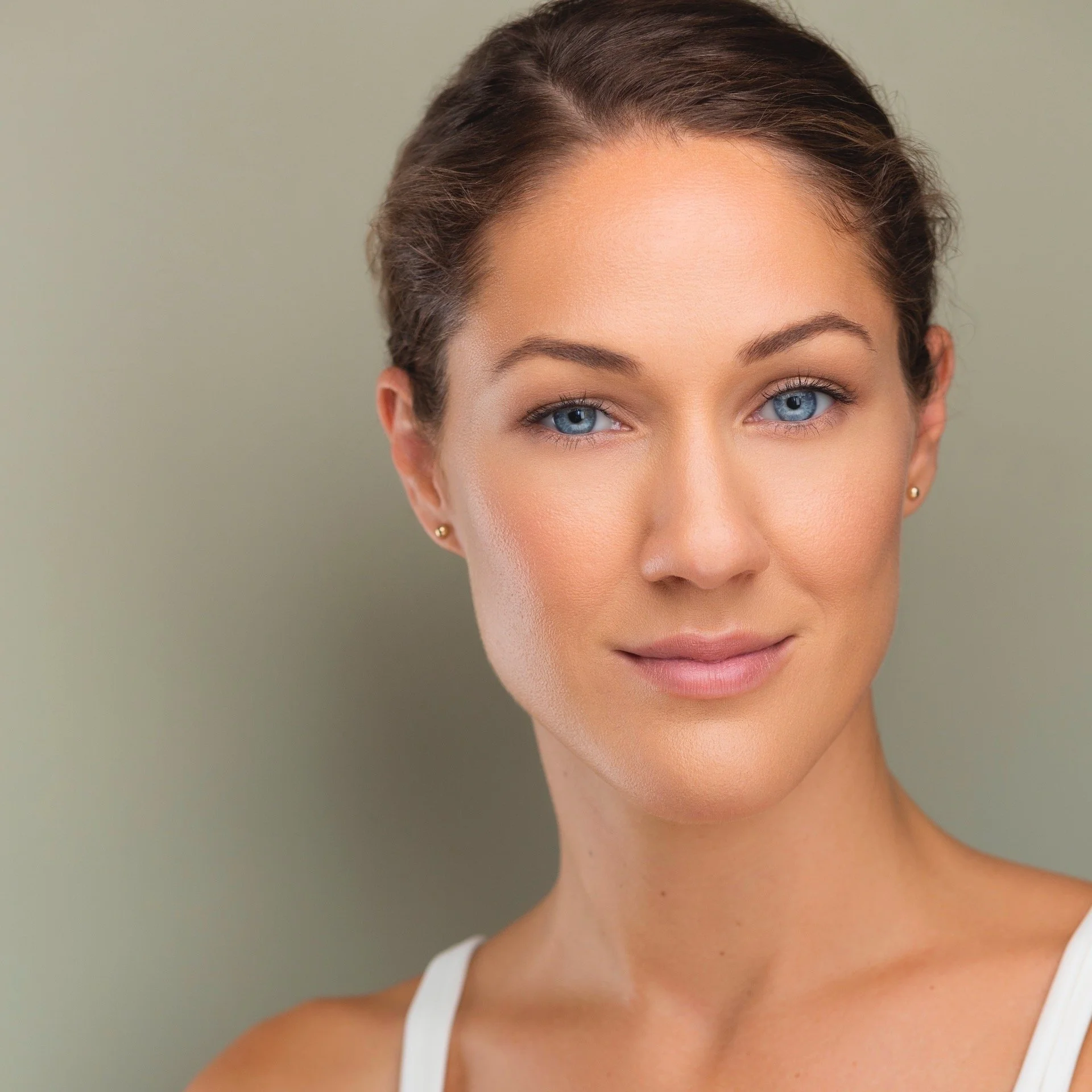 Close-up of a young woman with fair skin, blue eyes, brown hair styled back, and wearing a white tank top, smiling gently against a neutral background.