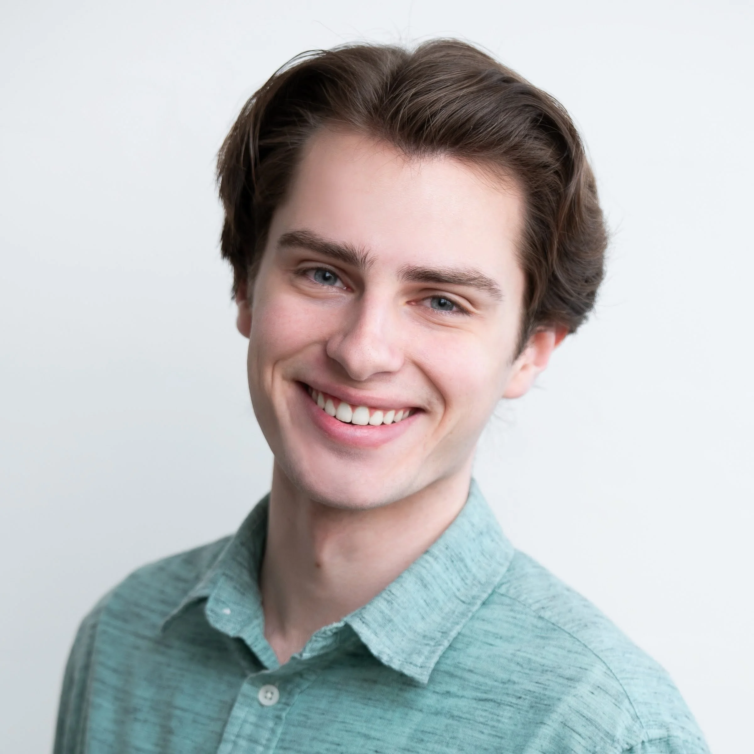 A young man with brown hair, blue eyes, and a bright smile, wearing a light green collared shirt, against a plain white background.
