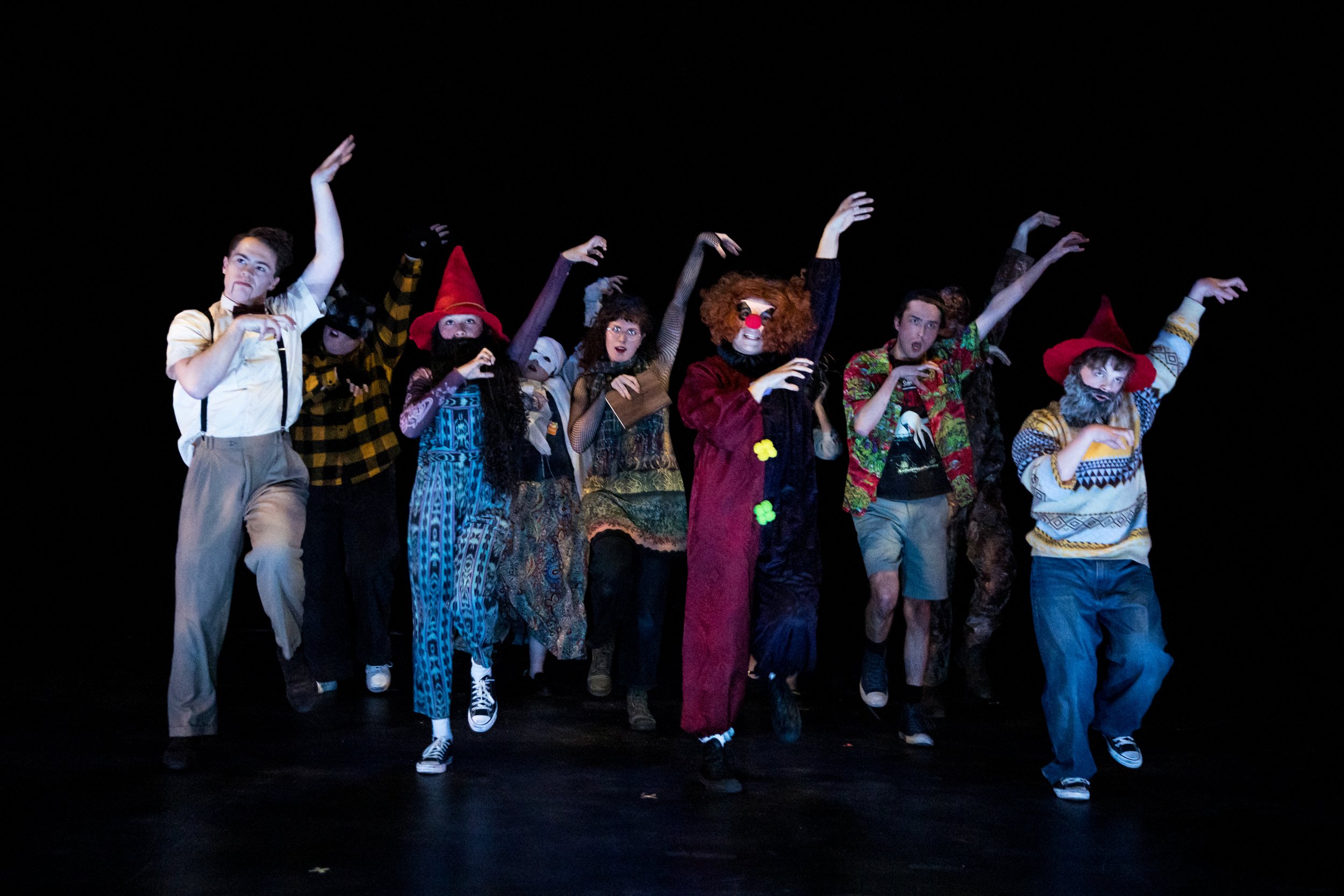 Theatre troupe performing on stage, dressed in eclectic costumes including clowns, witches, and casual wear, with a black background.