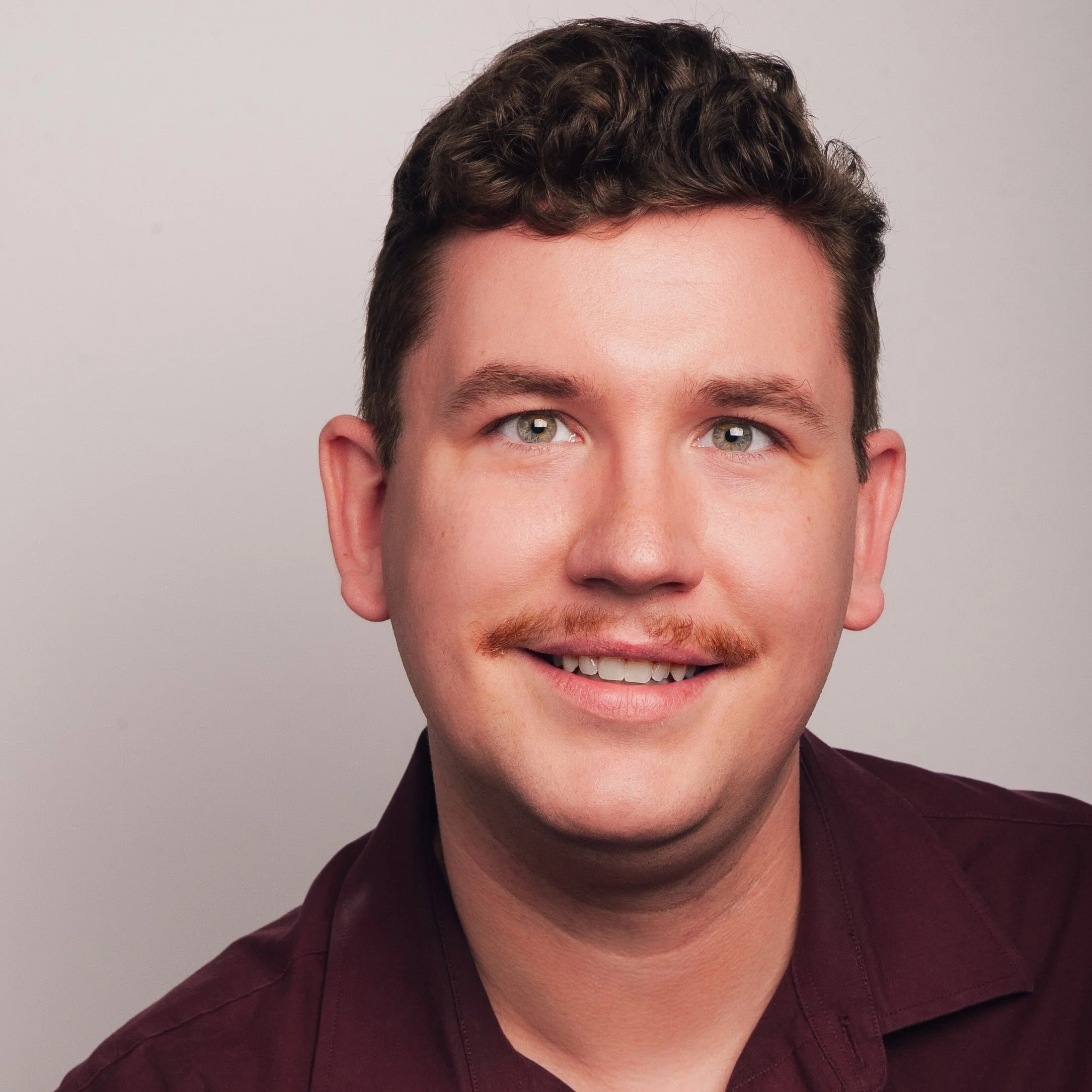 Close-up of a smiling young man with light skin, light blue eyes, short curly brown hair, and a mustache, wearing a dark red shirt, against a plain light gray background.