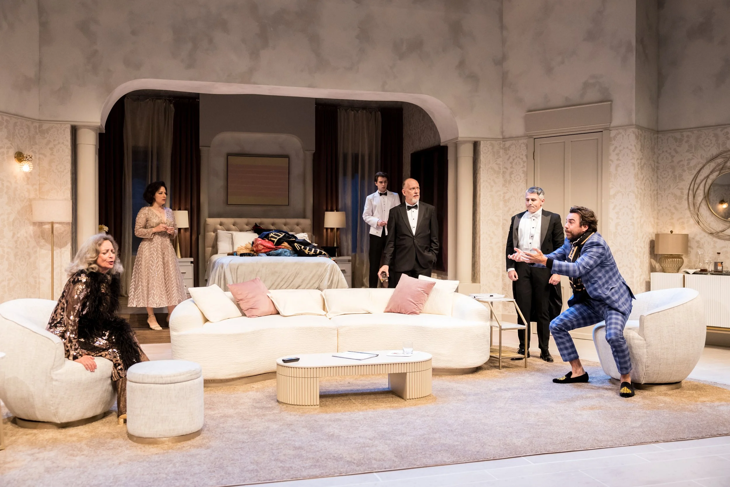 Six people in a living room scene with a bed in the background. One person seated on a stool, two people standing and engaging in conversation, one person in a blue checkered suit squatting and gesturing, one person in a tuxedo talking, and one woman