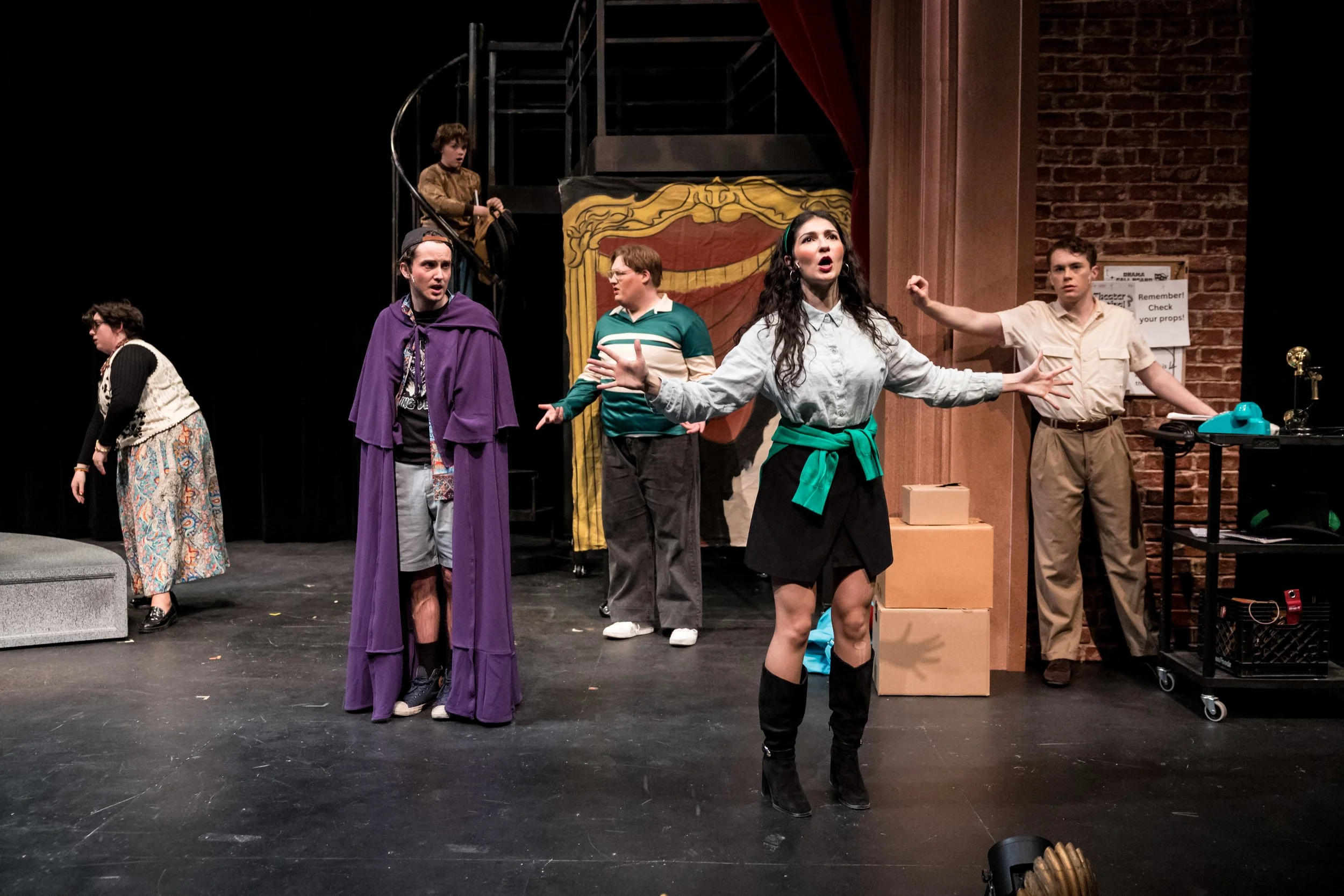 Actors performing on a stage with a brick wall backdrop, some standing and some with animated expressions, suggesting a theatrical scene.