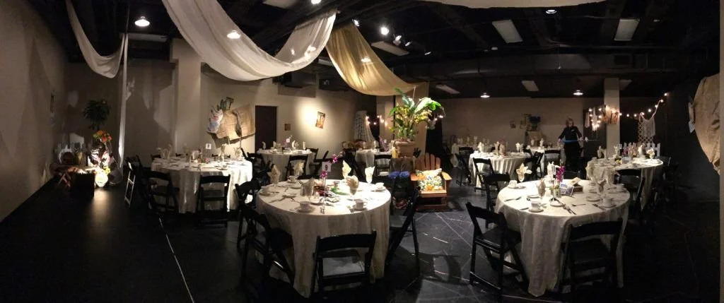 Setup of an indoor event space with round tables covered in white tablecloths, decorated with flowers and table settings. Draped fabric hangings and string lights are hanging from the ceiling, creating a festive atmosphere. There are some chairs posi