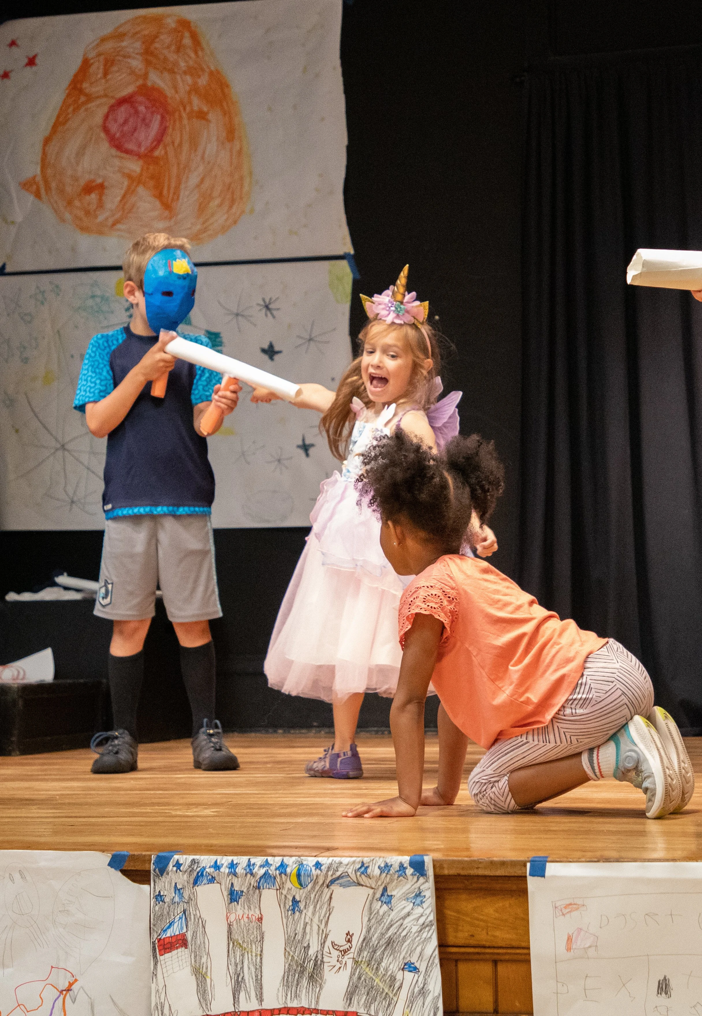 Children performing a play on stage, with one girl dressed as a unicorn and two boys wearing masks. One boy is holding a white paper sword, and a girl is on her knees, reacting emotionally.