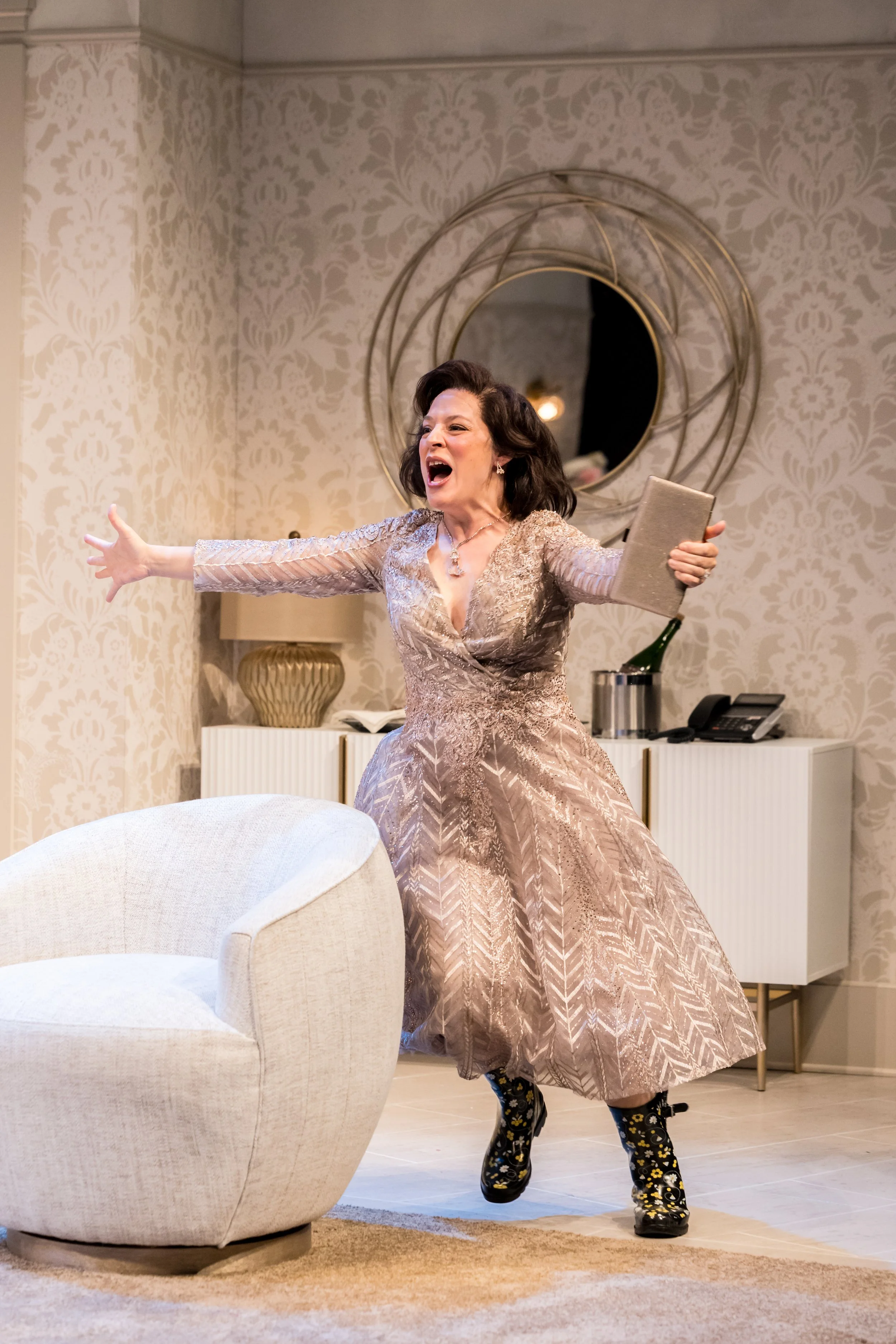 Woman in a glittering, long gown with floral patterned boots, standing inside a living room, gesturing passionately with a woman’s expression.
