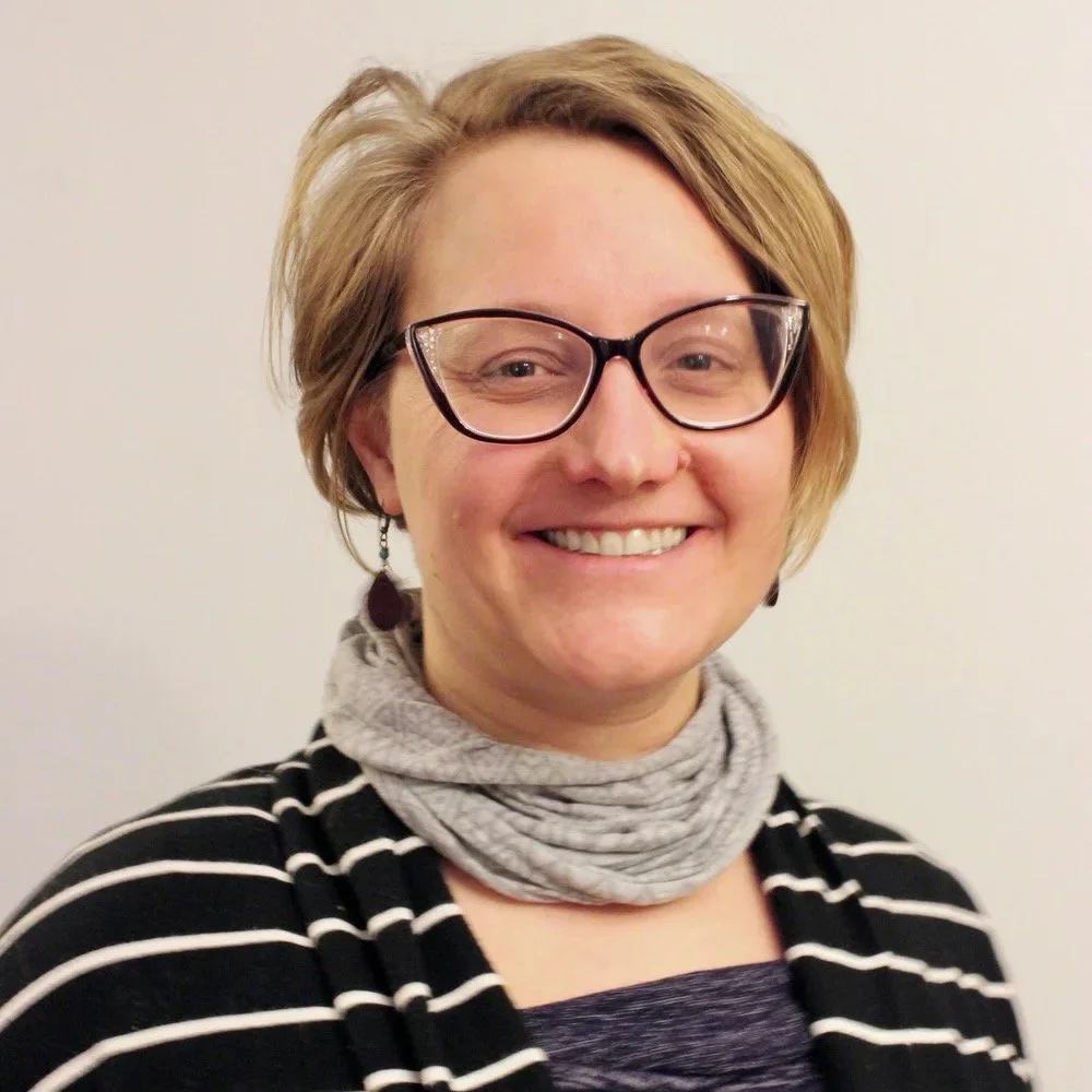 A woman with short, light brown hair wearing glasses, earrings, a gray scarf, and a black and white striped top, smiling in front of a plain white background.