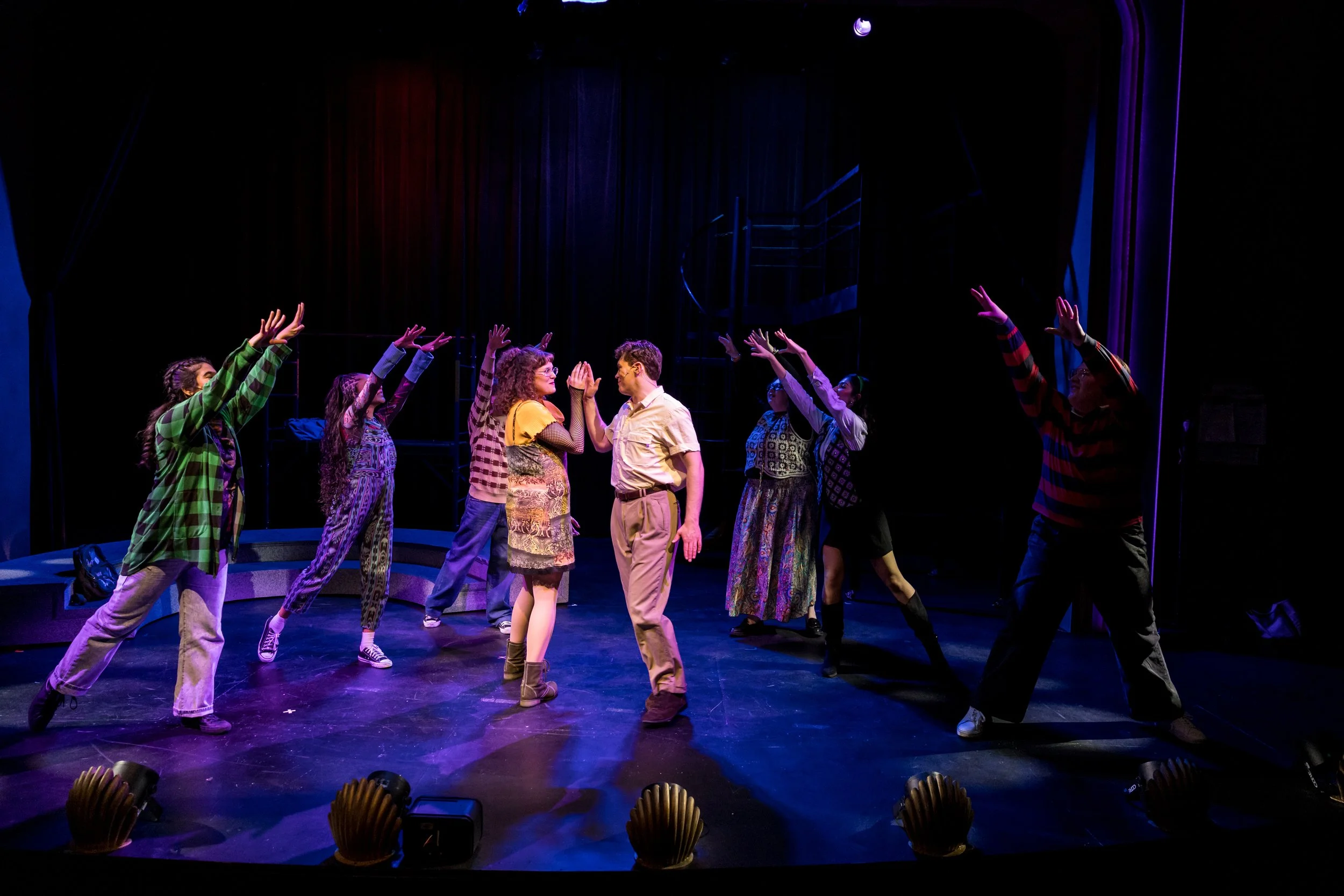 A theatrical stage performance with actors in casual clothing, engaging in a scene with many performers raising their hands, two actors in the center sharing a moment, under stage lighting with a dark background and red curtains.