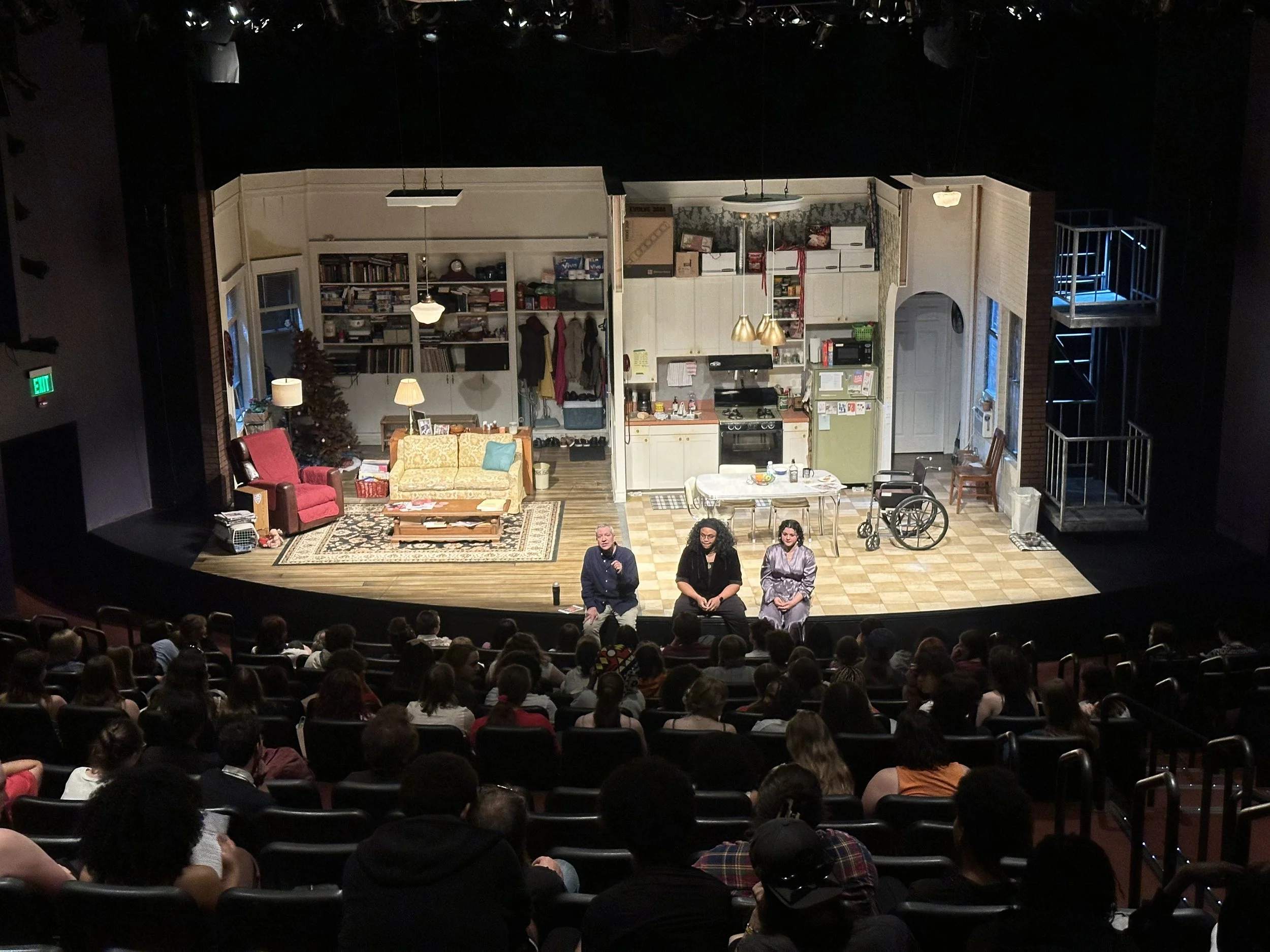 Theatrical stage set of a cozy living room and kitchen with cast members seated in front of an audience.