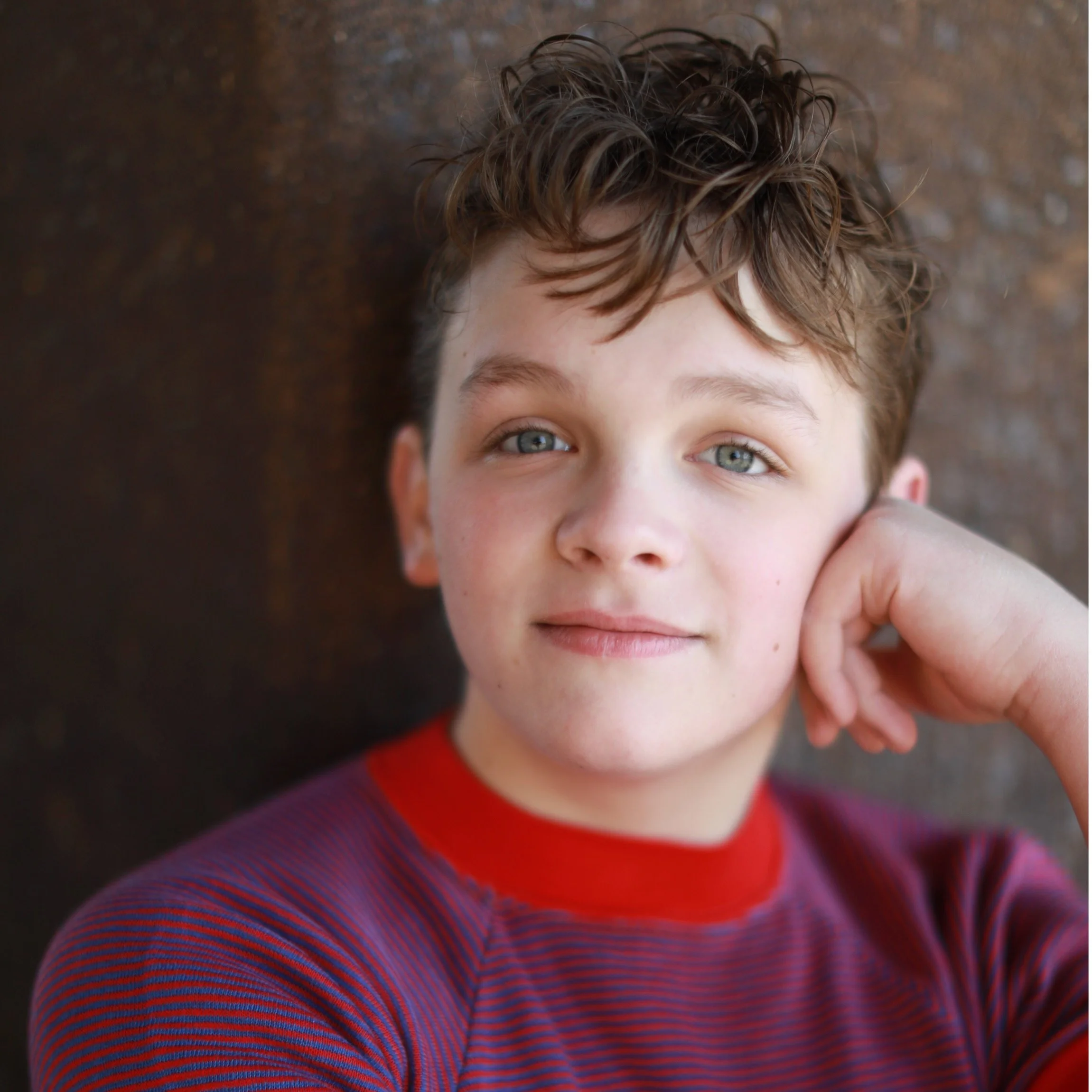 Close-up portrait of a young boy with curly brown hair and blue eyes, resting his head on his hand, looking at the camera with a slight smile.