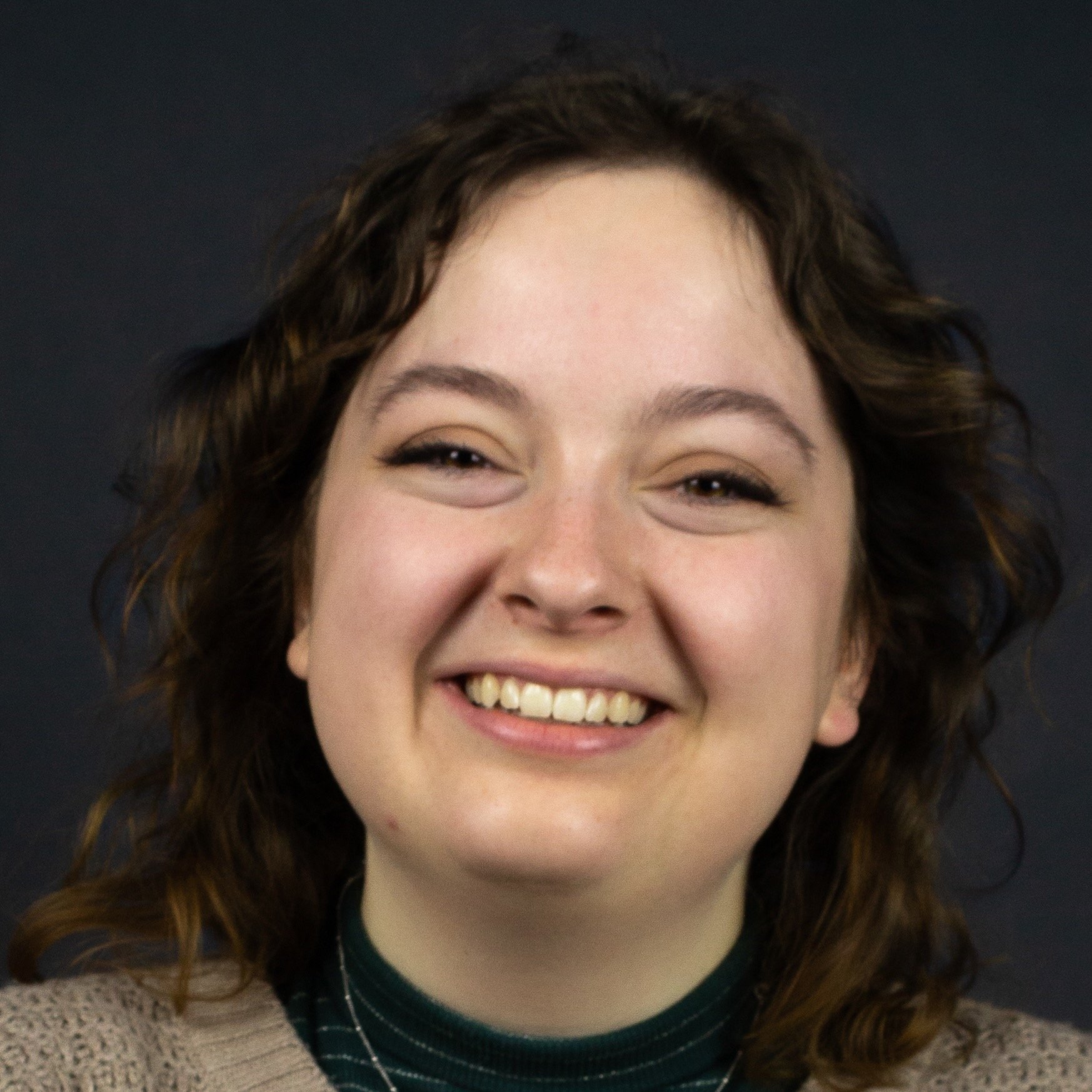 Close-up of a smiling woman with curly brown hair wearing a green turtleneck and beige sweater against a dark background.