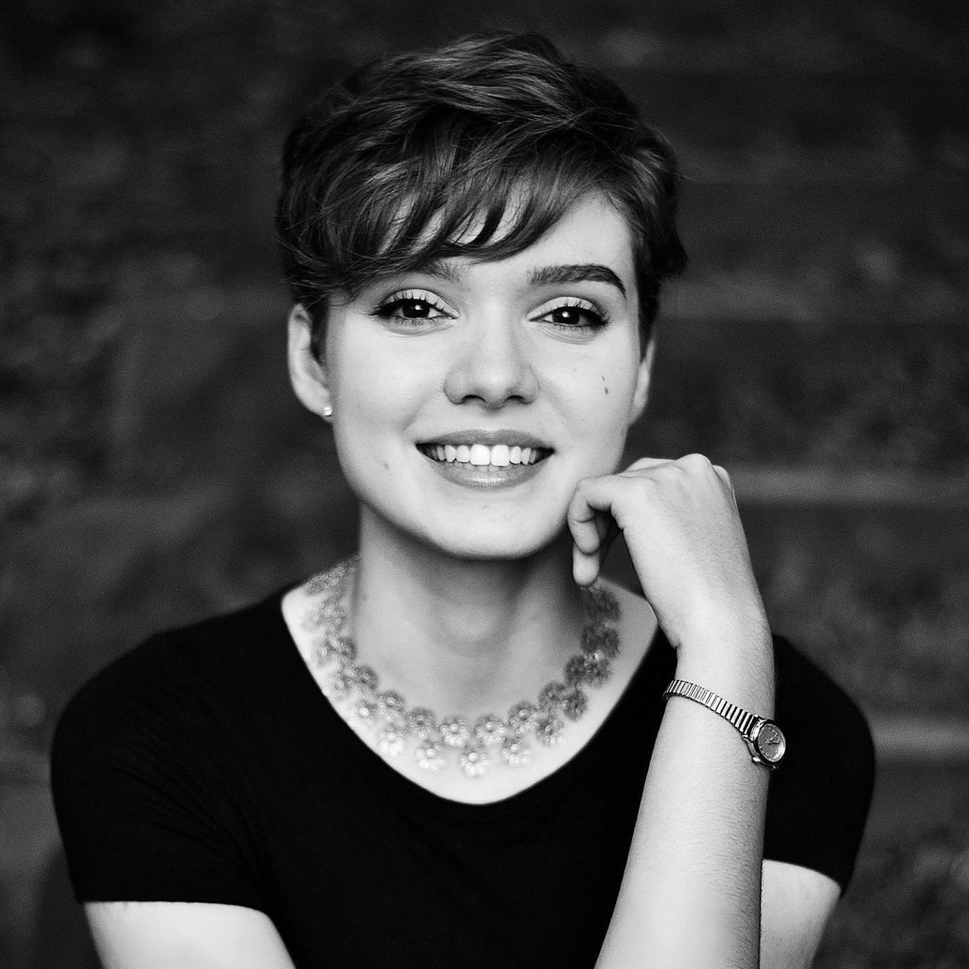 A smiling woman with short hair, wearing a necklace and a watch, resting her chin on her hand in a black and white photo.