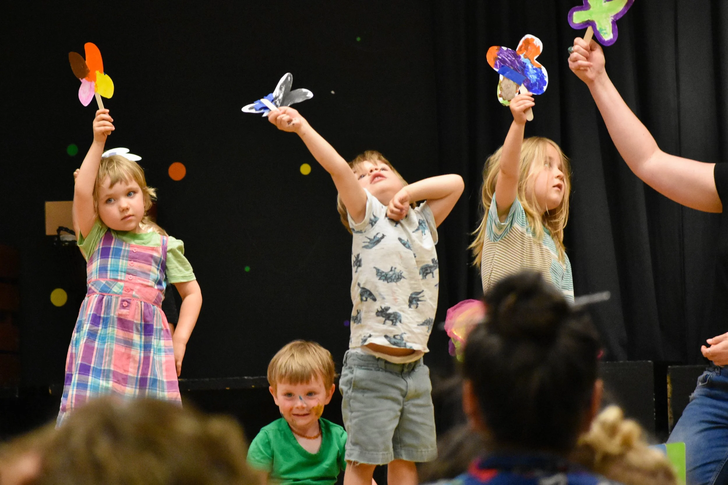 Children performing on stage holding colorful paper cutout props with a black curtain backdrop and colorful dot decorations.
