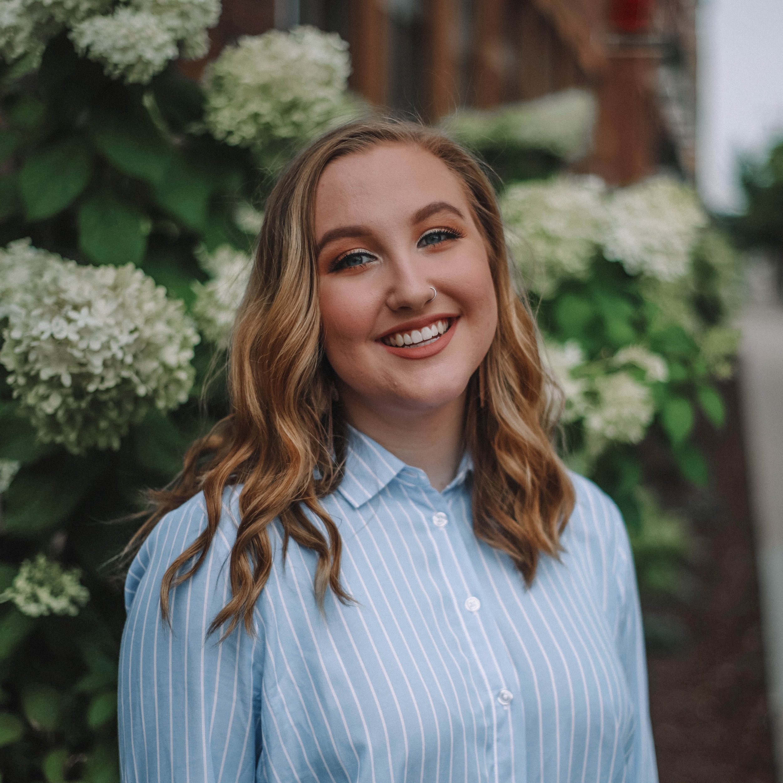 A smiling young woman with wavy blonde hair and a nose ring, wearing a light blue striped shirt, standing outdoors in front of white hydrangea flowers and green foliage.