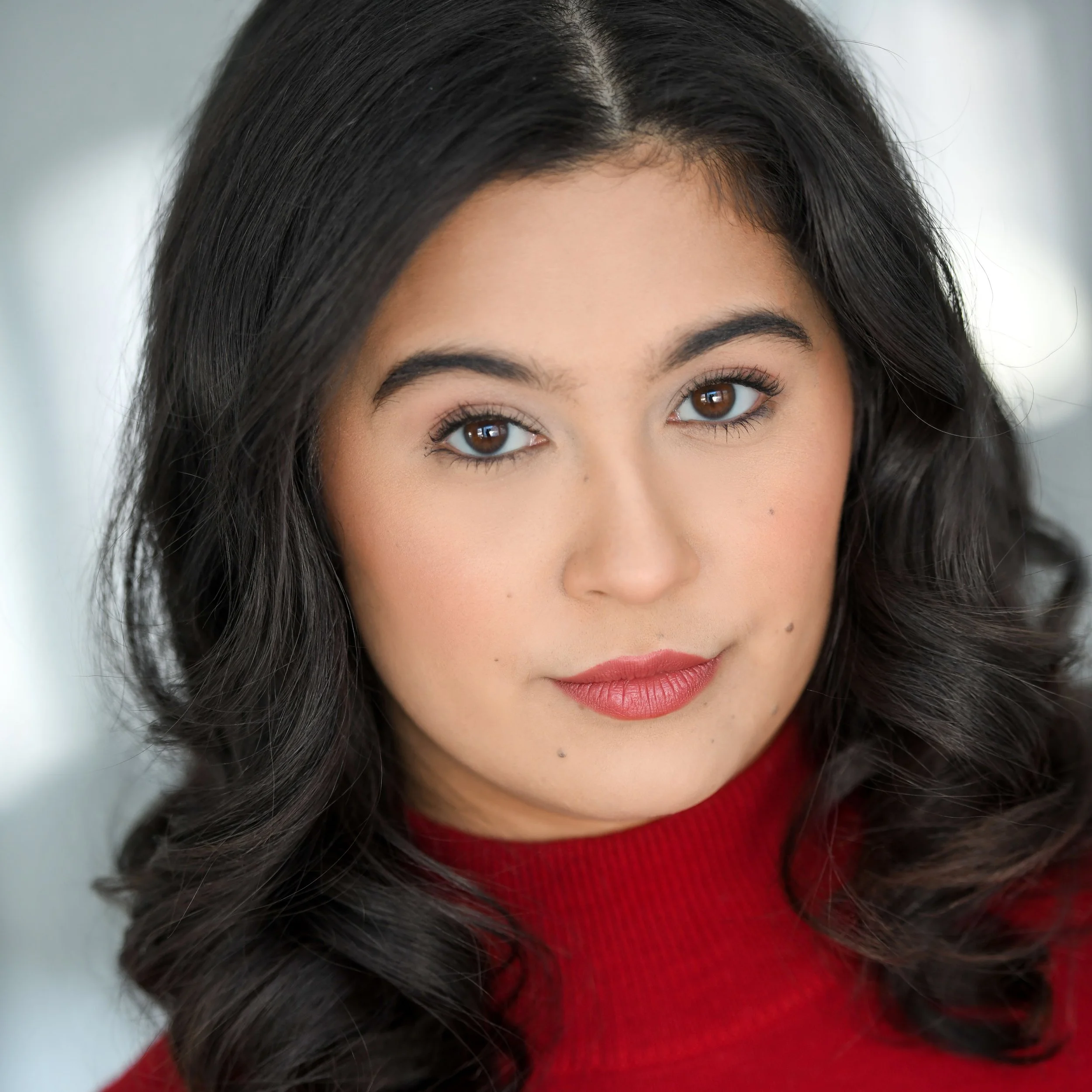 Close-up portrait of a young woman with dark, curly hair wearing a red top, looking directly at the camera.