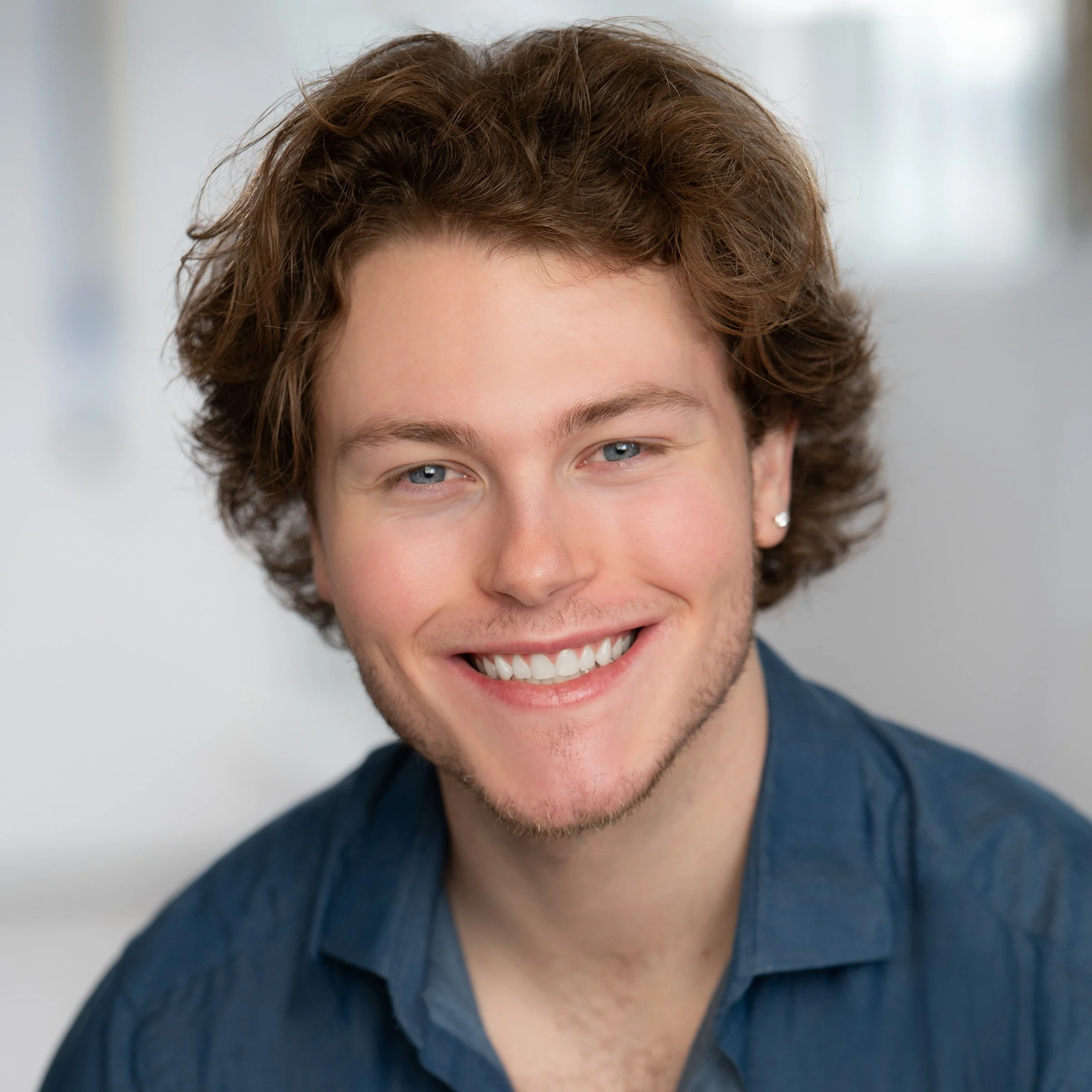 Close-up portrait of a young man with curly brown hair and blue eyes, smiling broadly, wearing a dark blue shirt.