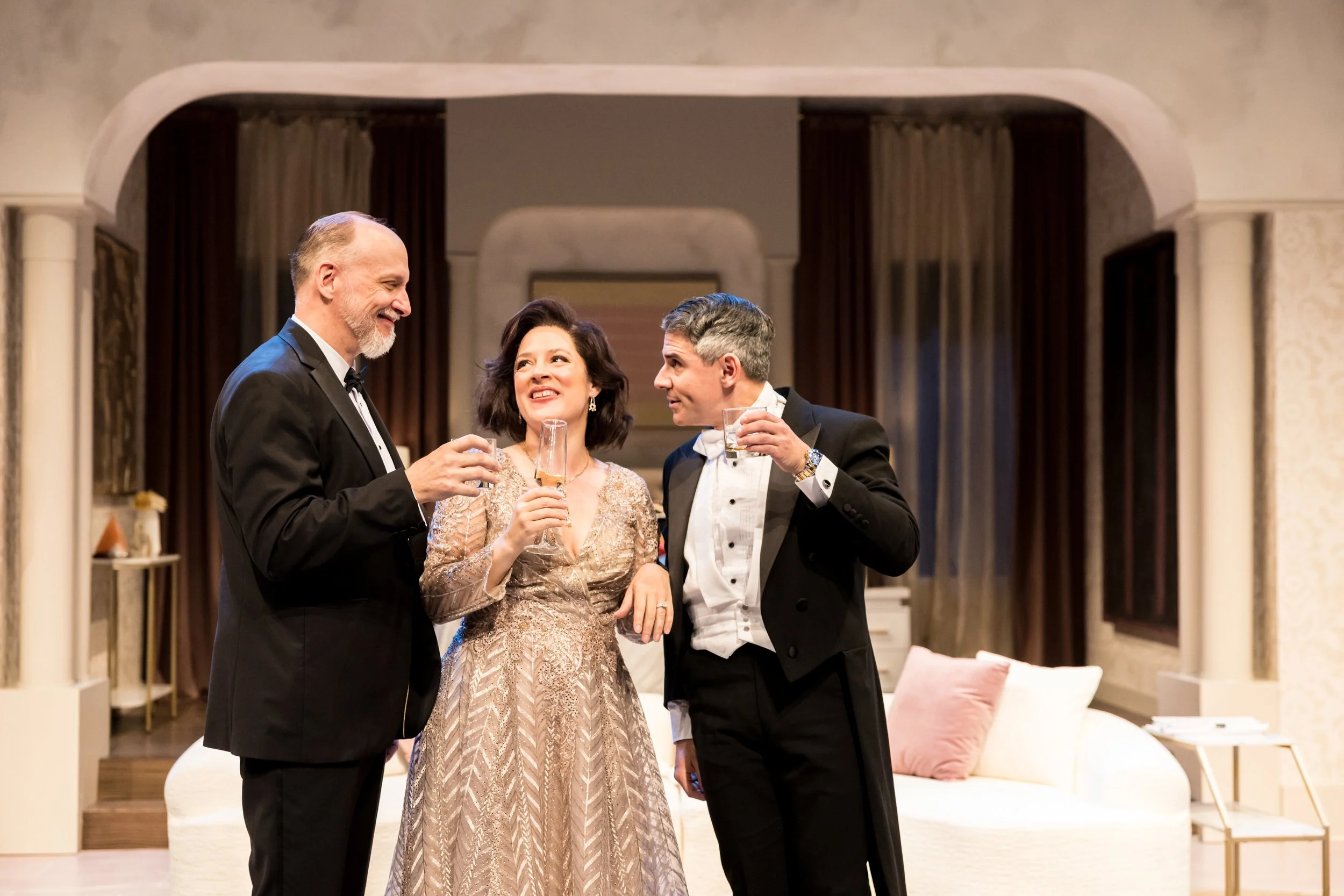 Three well-dressed people enjoying drinks and conversation in an elegant living room setting.