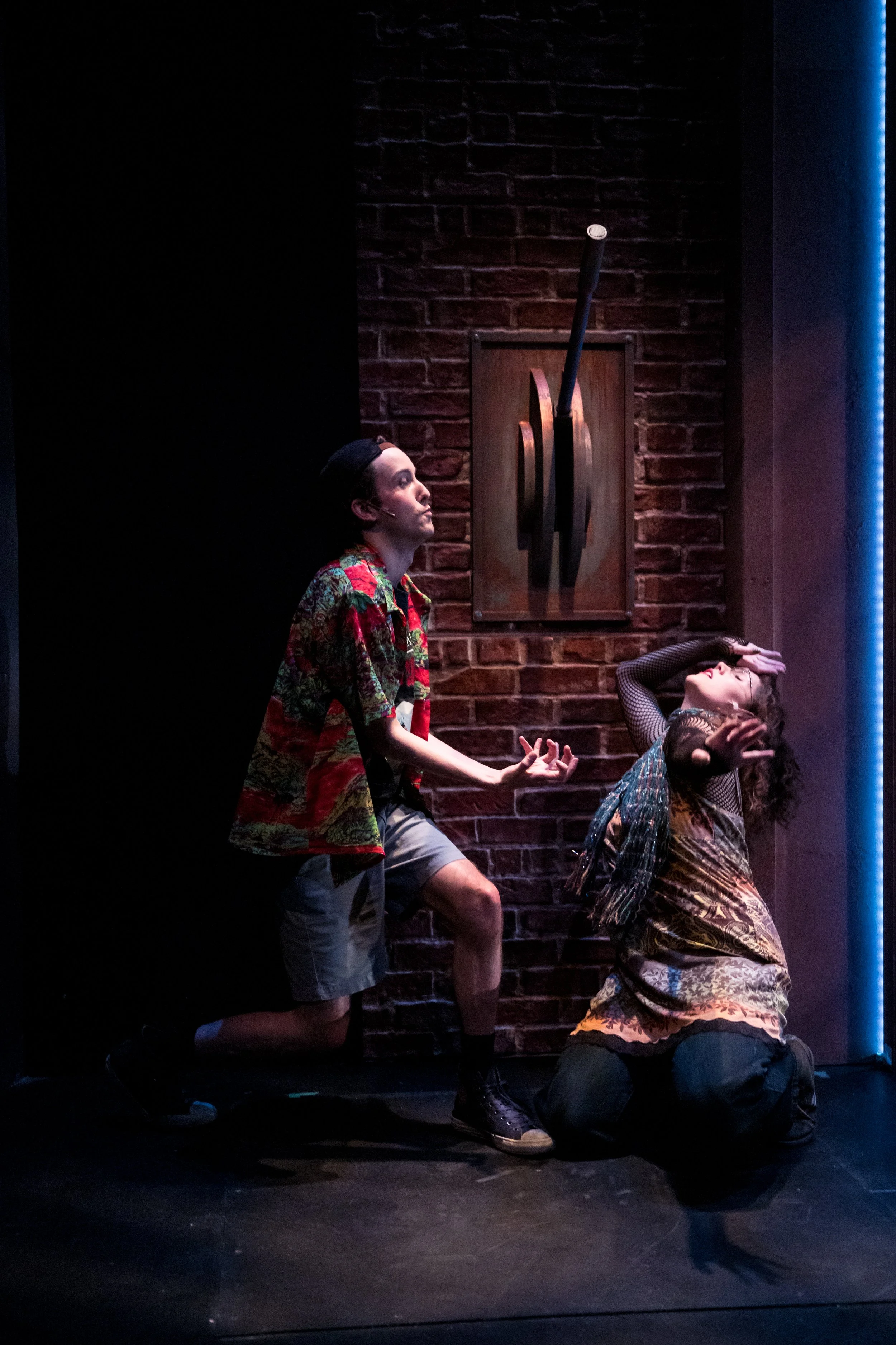 Two young performers on stage, one kneeling and the other standing, engaged in a dramatic scene against a brick wall backdrop with abstract art.