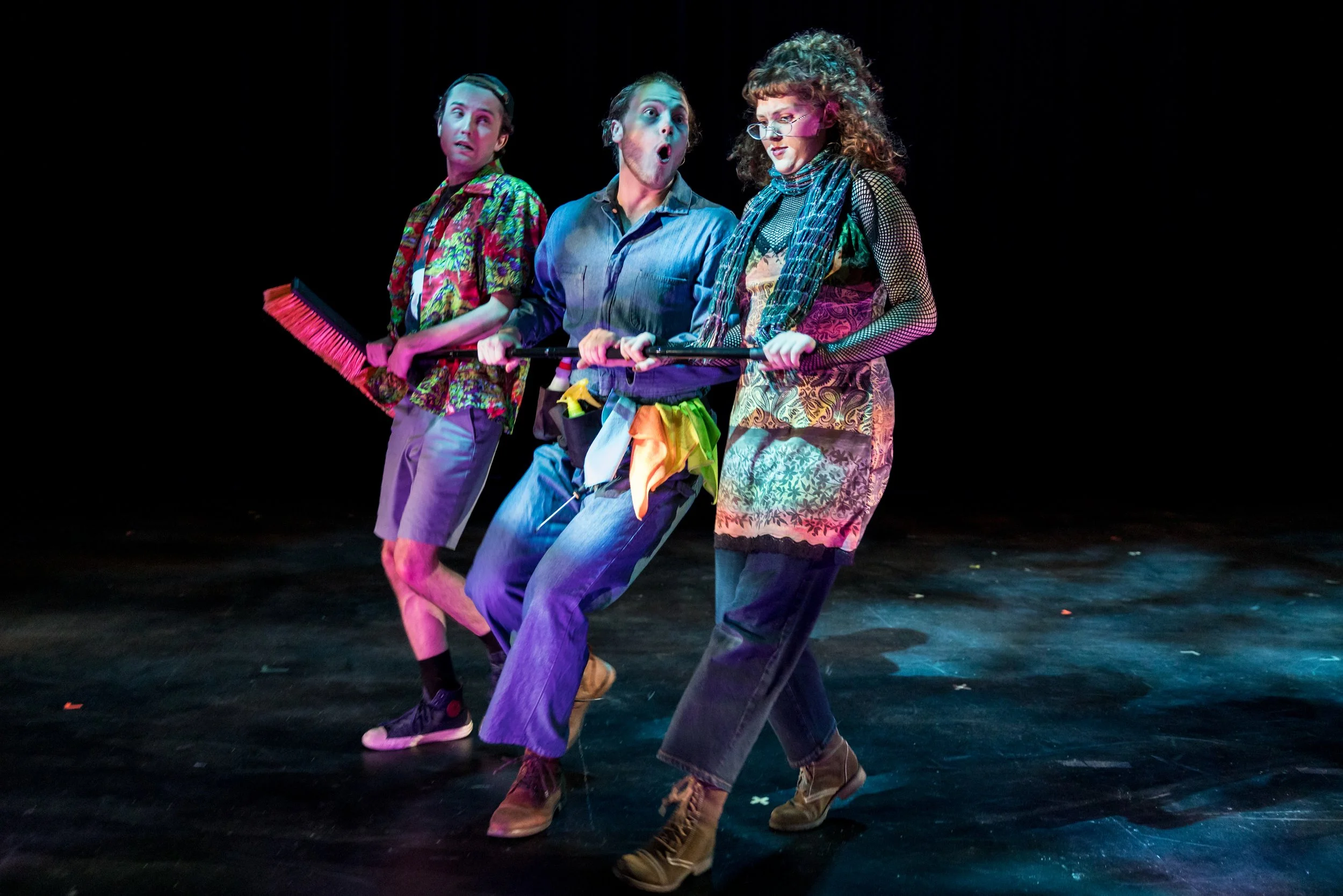 Three actors on stage, dressed in colorful, casual costumes, holding a broom, appearing surprised or scared during a theatrical performance.