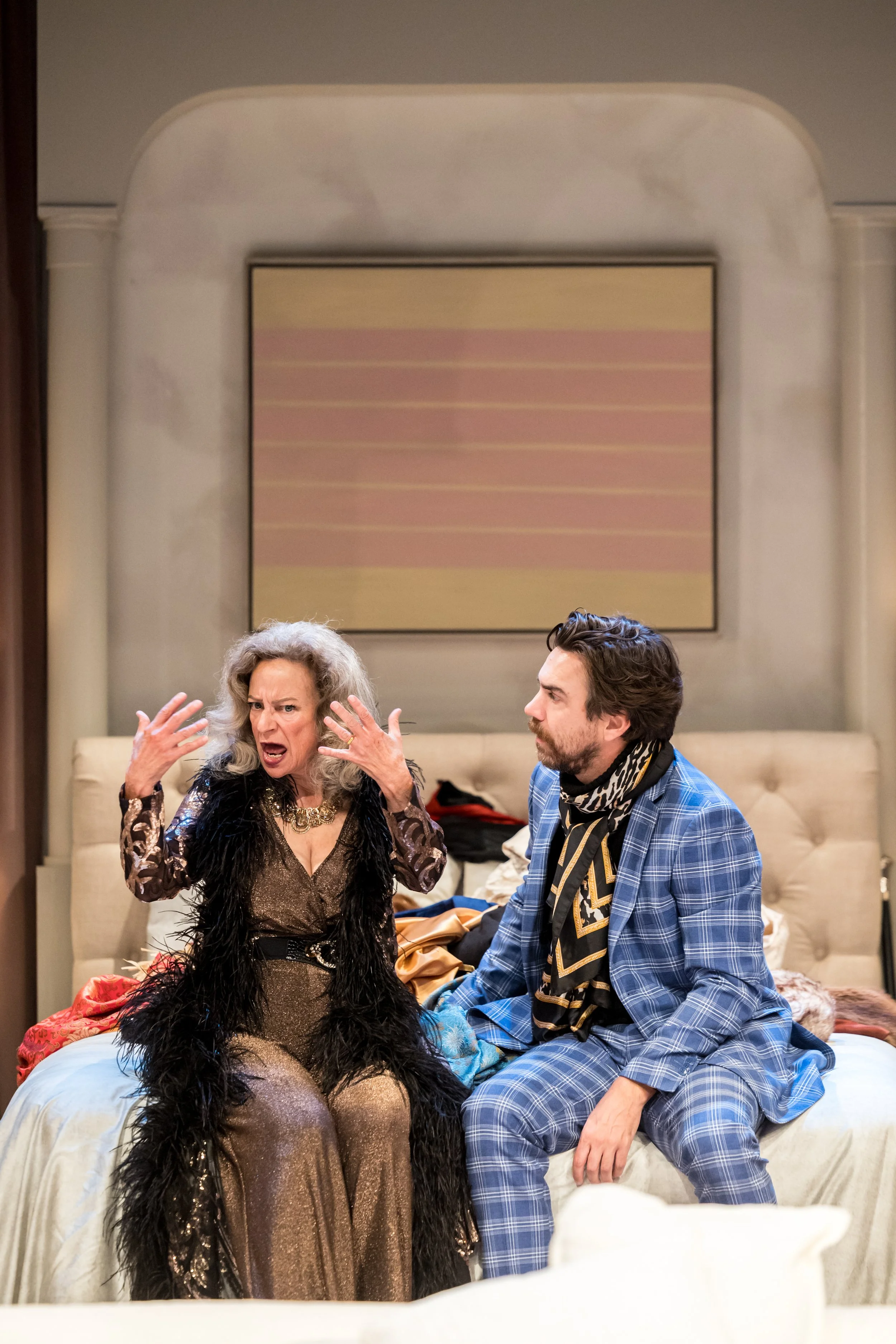 A woman with gray hair yelling and gesturing passionately while sitting on a bed next to a man with brown hair and beard, wearing a blue plaid suit and scarf. The scene appears to be from a theatrical performance or a TV show.