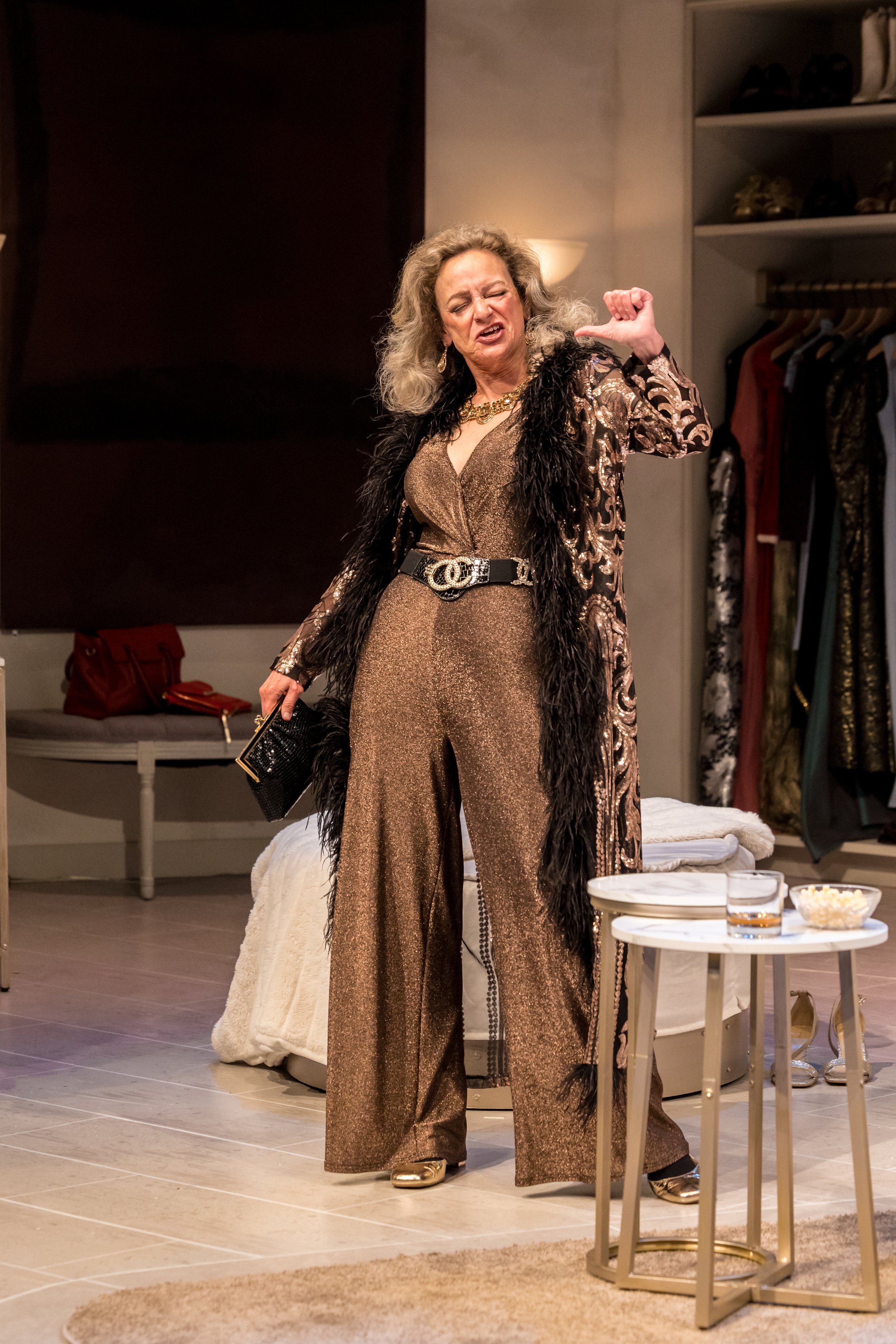 A woman dressed in a shiny, patterned jumpsuit with a black fur trim coat, gold shoes, gold jewelry, and a black belt, standing in a bedroom.