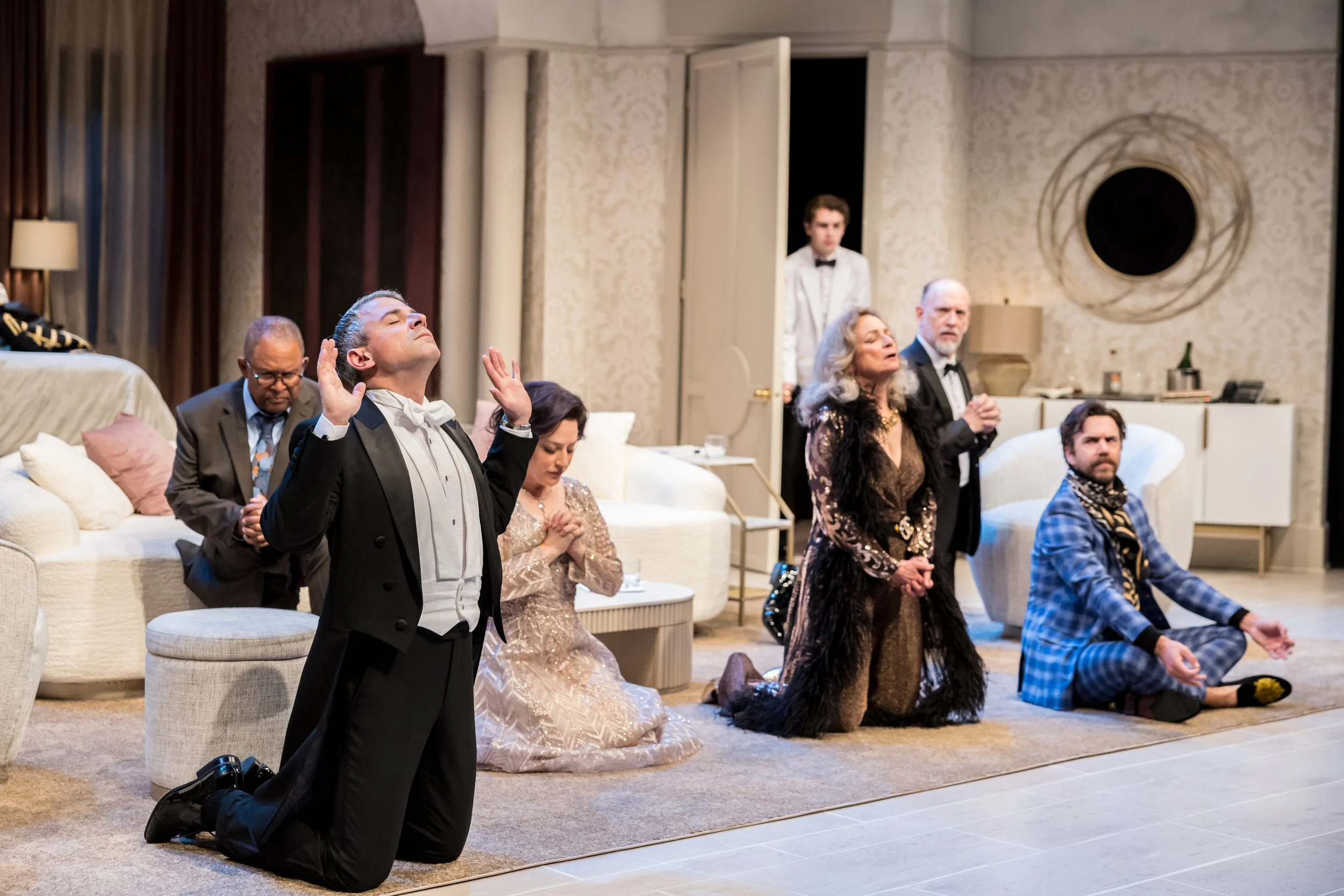 People dressed in formal and eccentric outfits kneeling with eyes closed, praying or meditating in a living room with beige wallpaper and furniture.