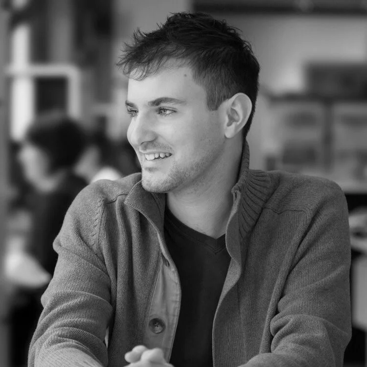 A smiling young man with short, dark hair, wearing a cardigan over a black shirt, sitting at a table in a social setting.