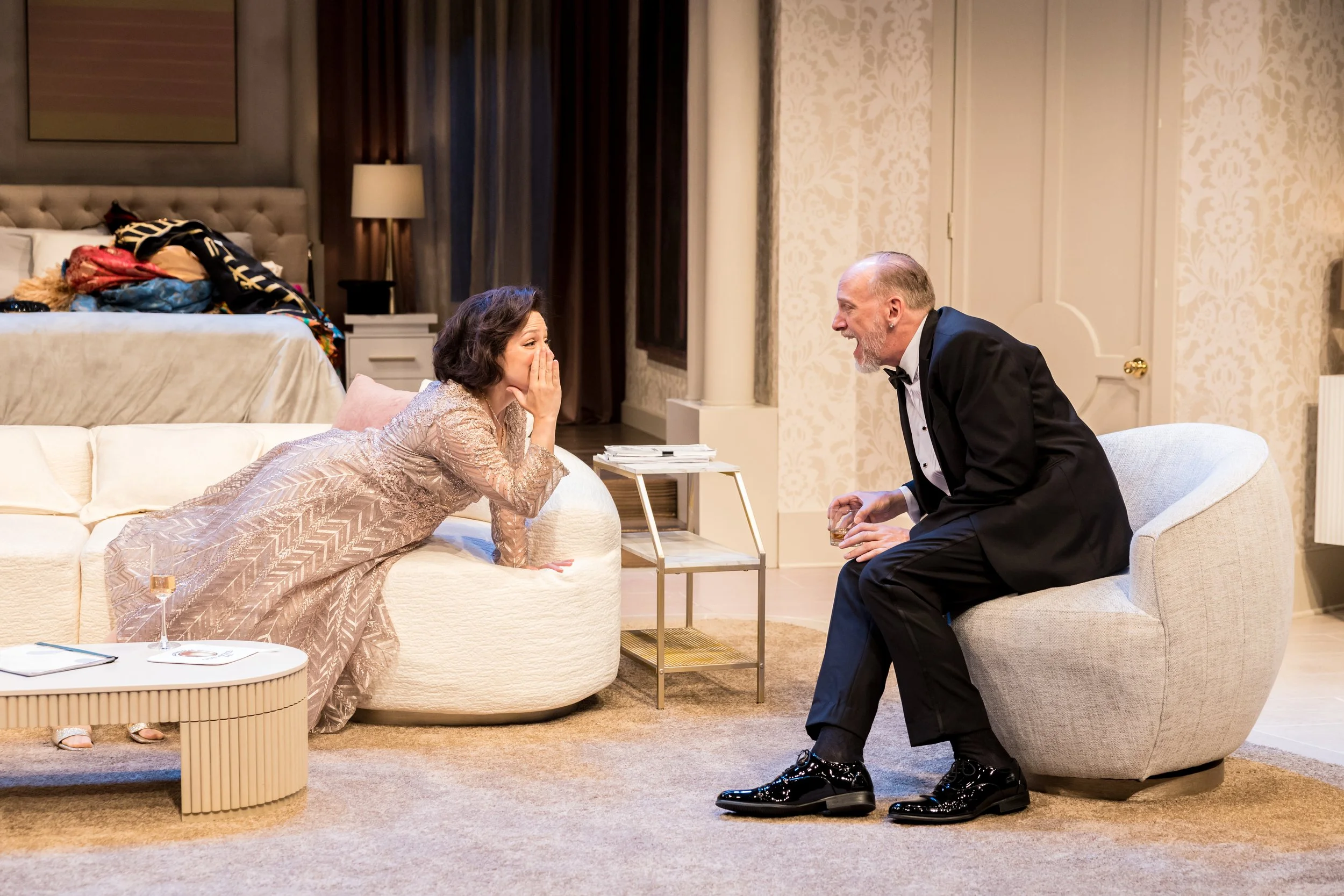 A woman in a beige dress is sitting on a white couch, covering her mouth with her hand, smiling. A man in a black tuxedo is sitting on an armchair, holding a glass, smiling at her. The room is a decorated hotel suite with a bed, side tables, and a pi