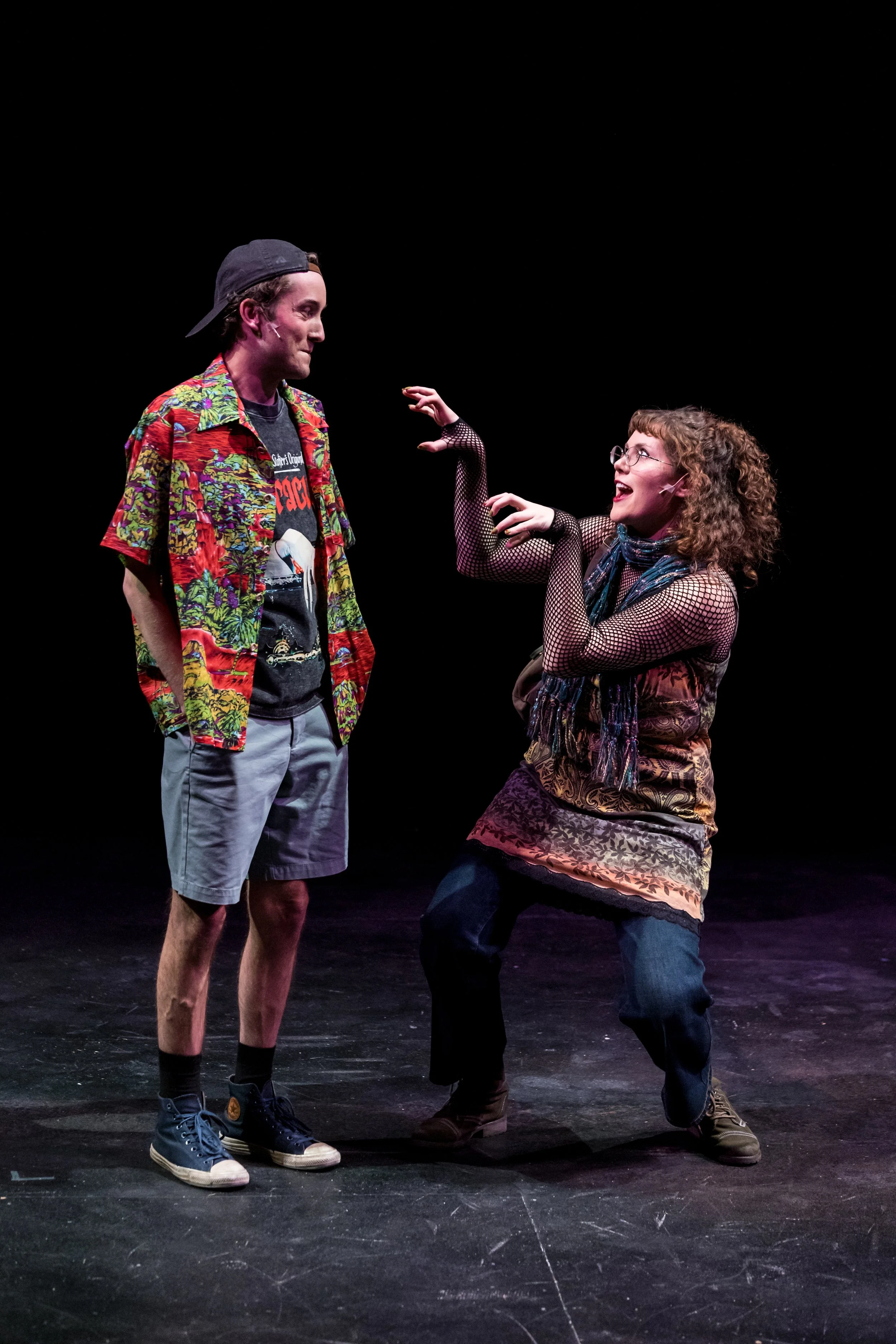 Two actors performing on stage, one man wearing a colorful Hawaiian shirt, gray shorts, and a backward cap, and a woman wearing glasses, a patterned dress, fishnet sleeves, and a colorful scarf, engaged in a theatrical scene.