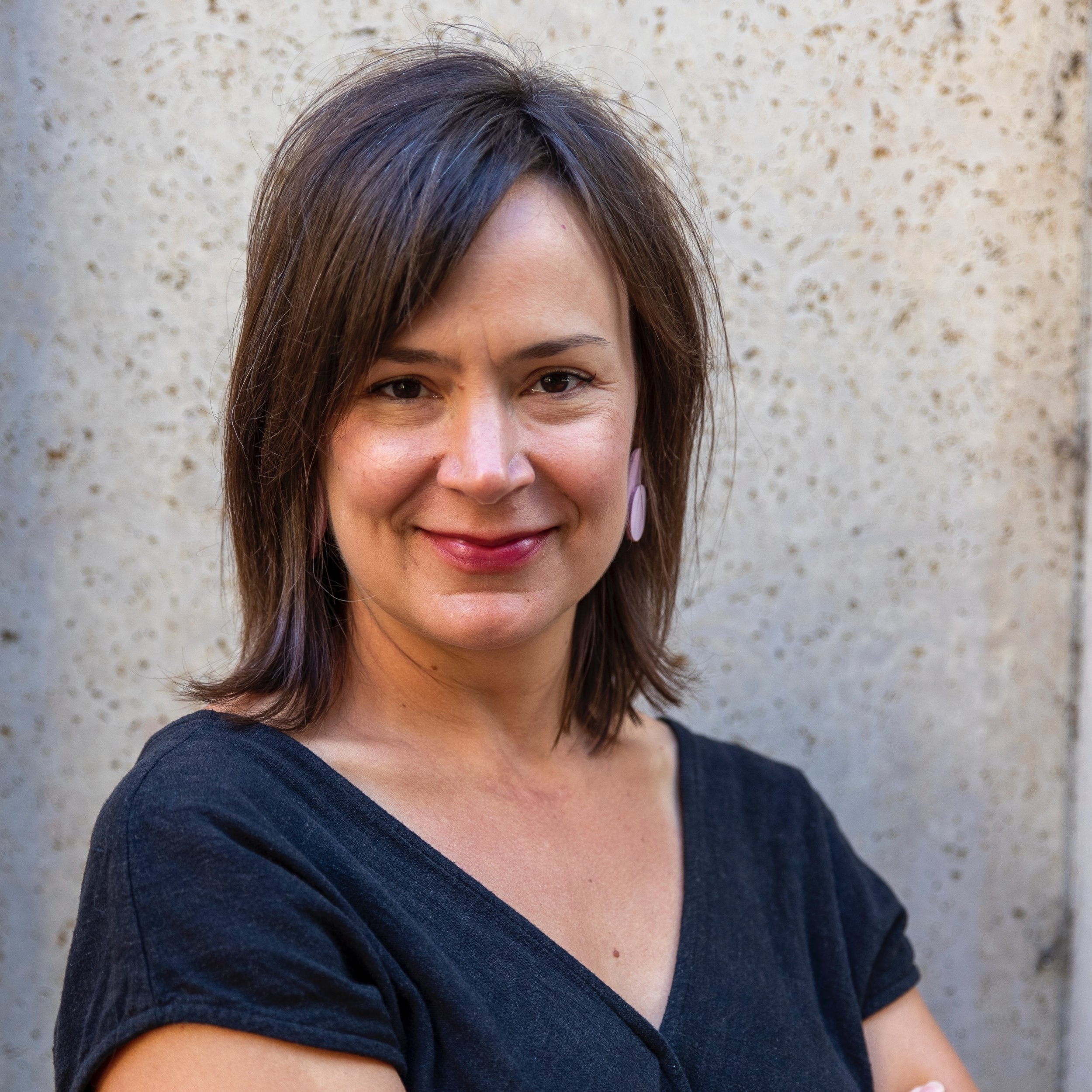A woman with shoulder-length brown hair, wearing a black top and pink earrings, standing against a textured beige wall.