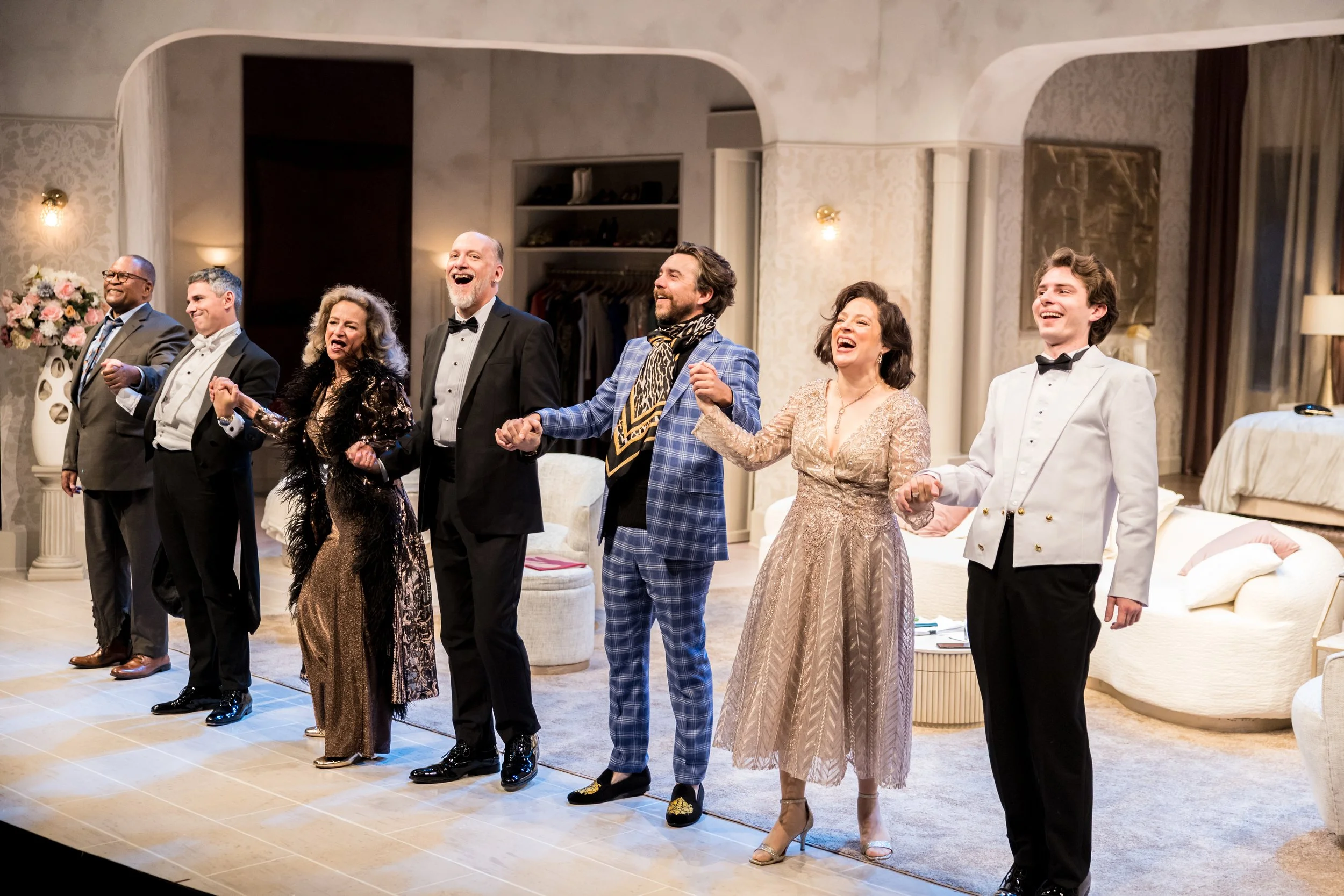 A group of seven people dressed in formal attire, holding hands, and appearing to be singing or celebrating in a well-lit, elegant room with floral arrangements and luxurious decor.