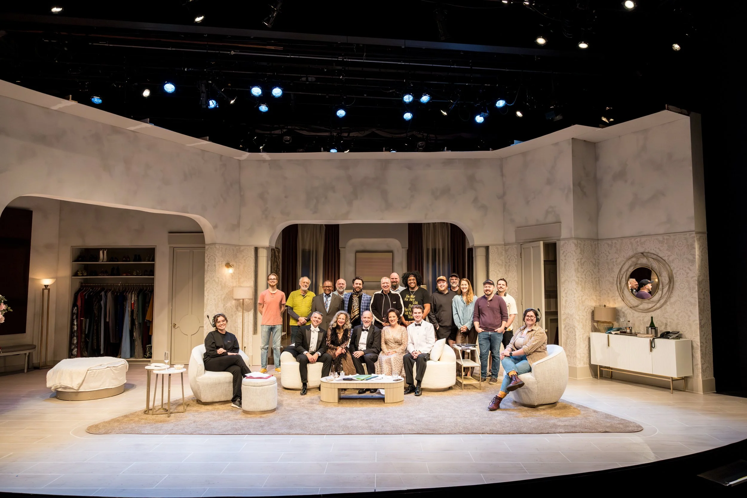 The cast of a theatrical play or TV show posed for a photo on stage, in a living room set with cream-colored walls, furniture, and decor. The group includes men and women, some seated on sofas and chairs, others standing behind. The stage is lit with
