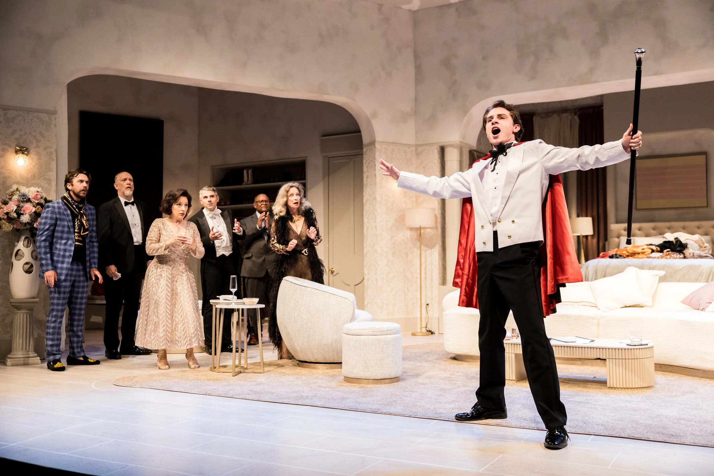 A theatrical performance featuring a man dressed as a prince singing or speaking passionately with arms outstretched in a fancy living room setting, while six people watch with various expressions.