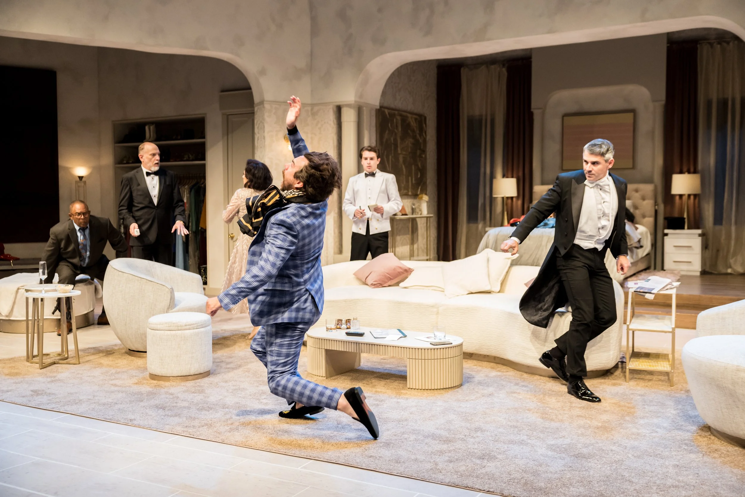 Dramatic scene from a theatrical play with actors in tuxedos and formal wear on stage, with some in motion and disarray, set in a luxurious living room.