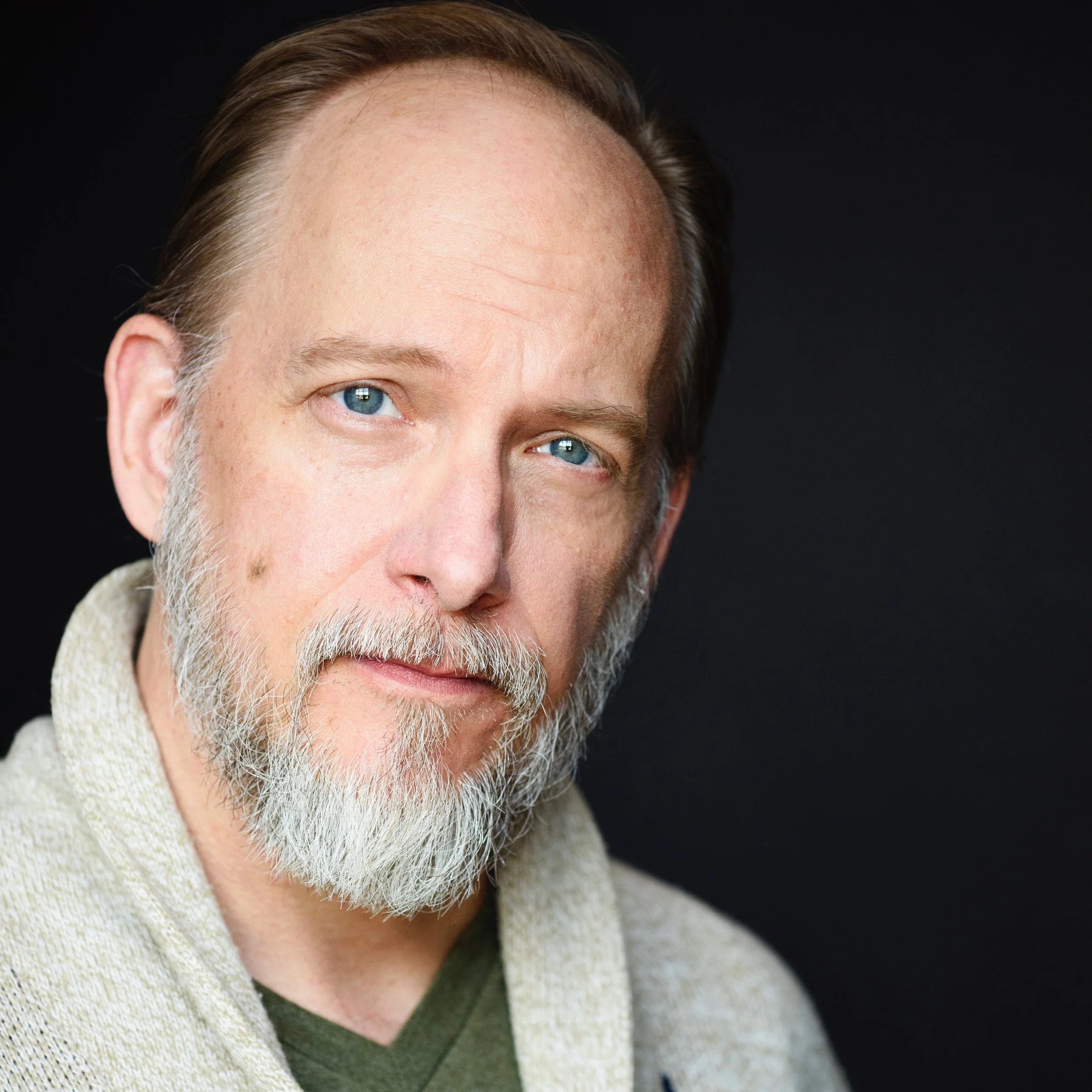 Headshot of middle-aged man with blue eyes, gray beard and mustache, wearing a light-colored cardigan over a dark green shirt, against a dark background.
