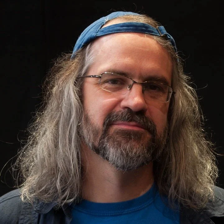 Portrait of a man with long gray hair, a beard, glasses, wearing a blue headband and a blue shirt, against a black background.