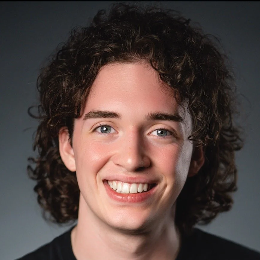 Close-up portrait of a young man with curly brown hair, blue eyes, and a big smile, wearing a black shirt.