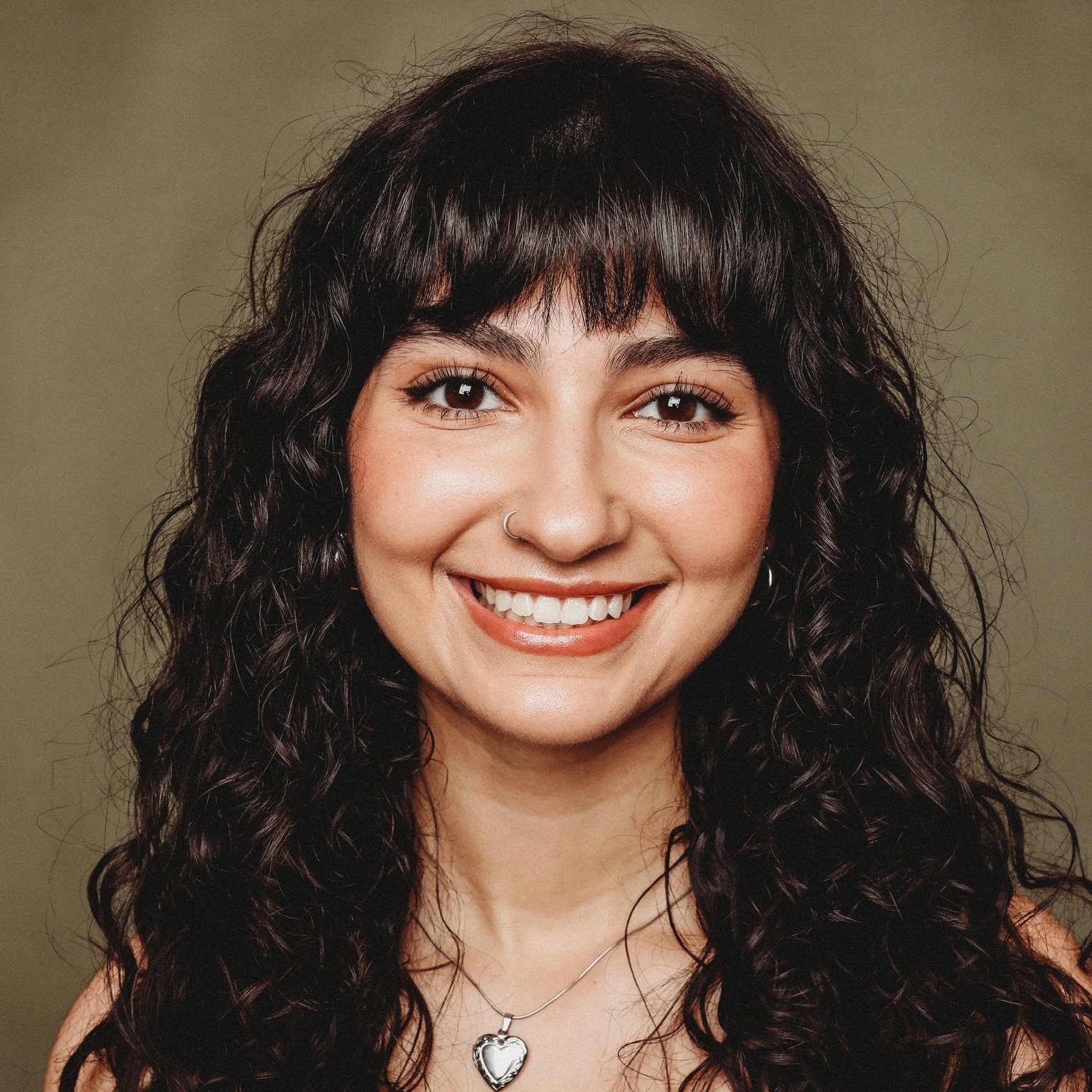 Close-up of a woman with dark, curly hair, a nose ring, and a heart-shaped pendant necklace smiling at the camera.
