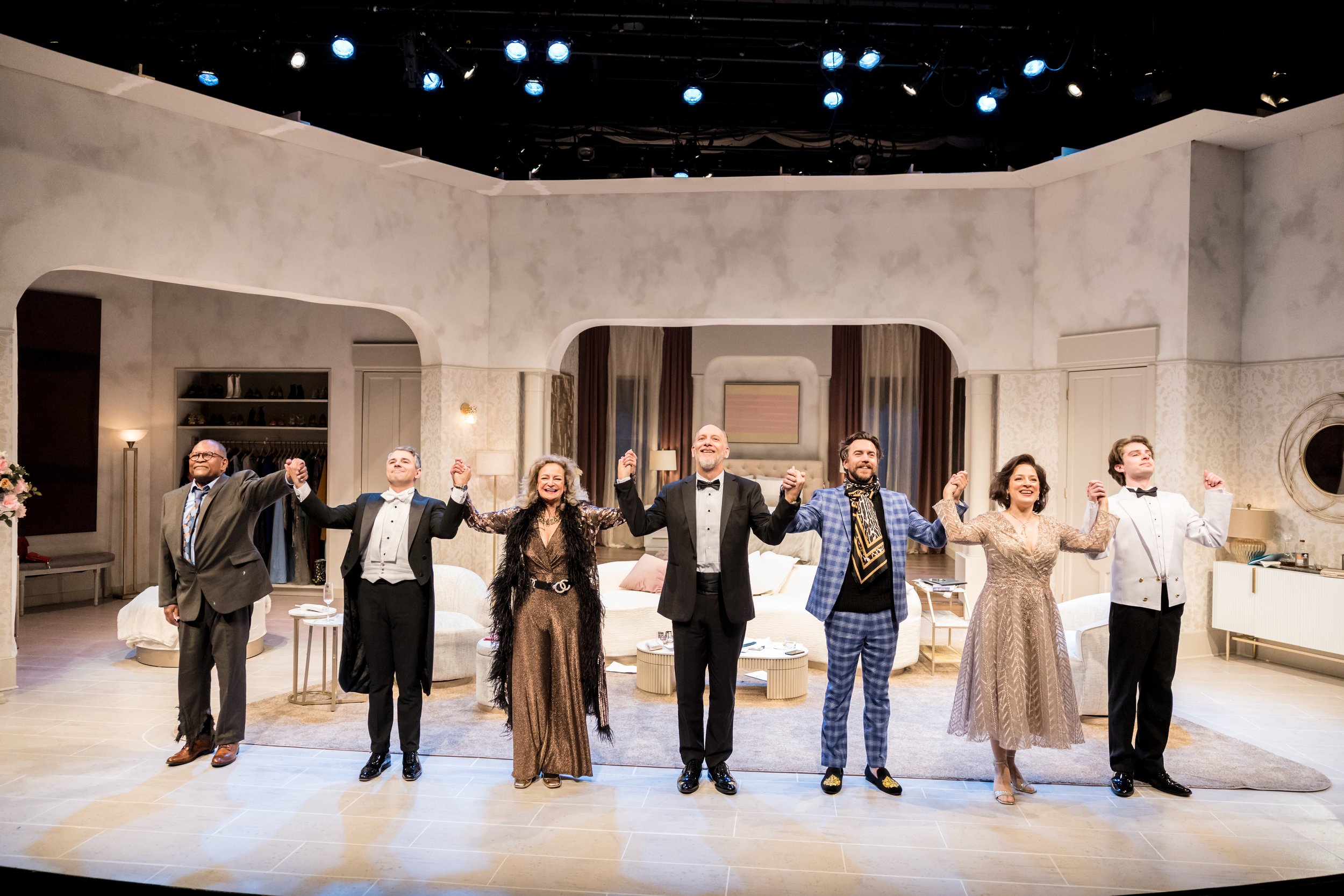 The cast of a theater play standing in a line holding hands and taking a bow on a set resembling a living room, with bright lights shining down.