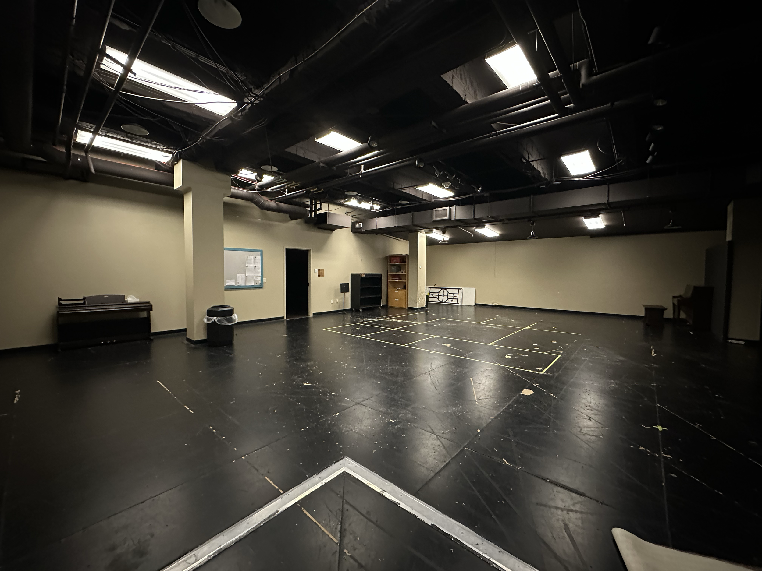 Empty black stage with some yellow tape markings and a few scattered pieces of debris in a room with beige walls, exposed ceiling pipes, and fluorescent lights.