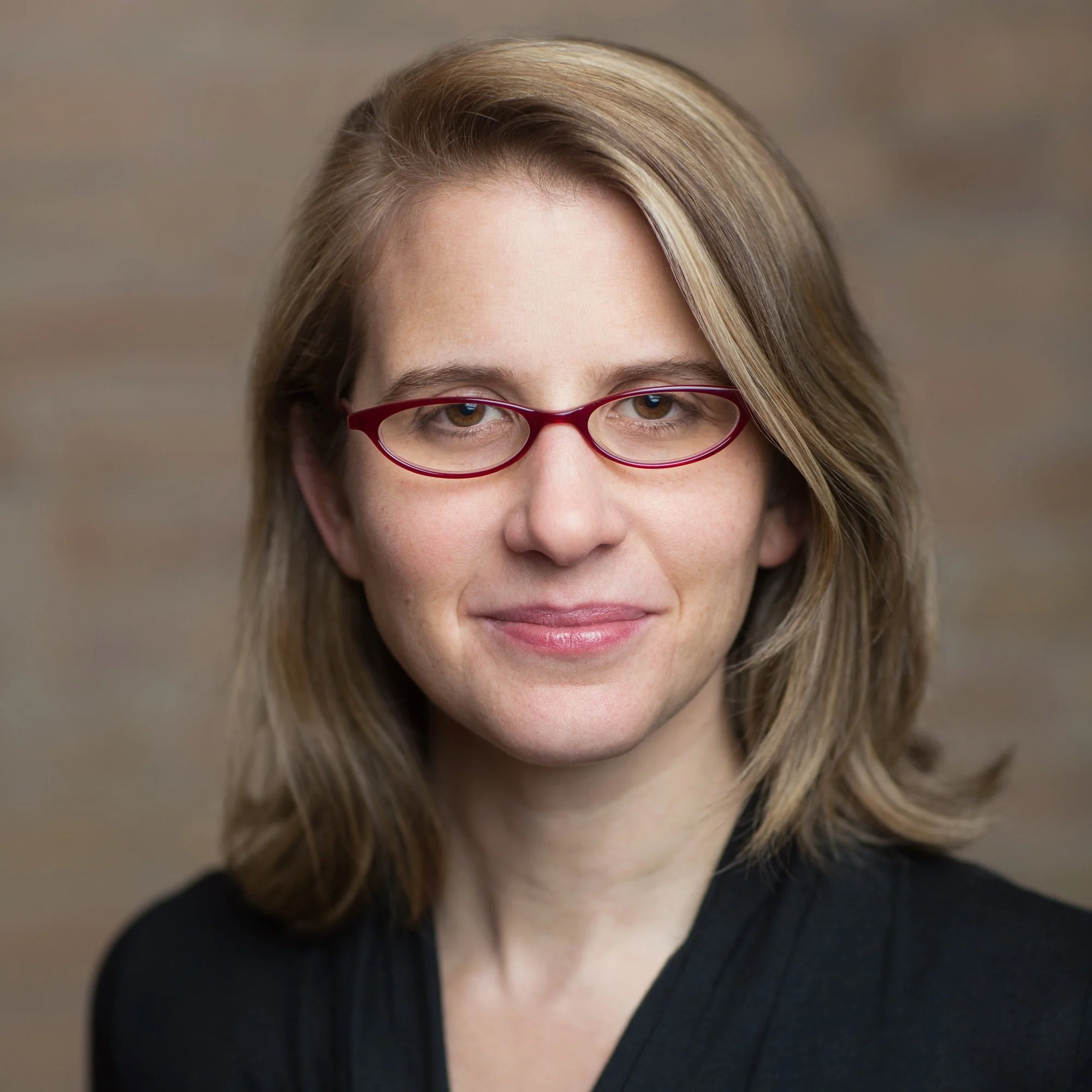 Close-up portrait of a woman with shoulder-length light brown hair, wearing red glasses and a black top, smiling softly at the camera against a neutral background.