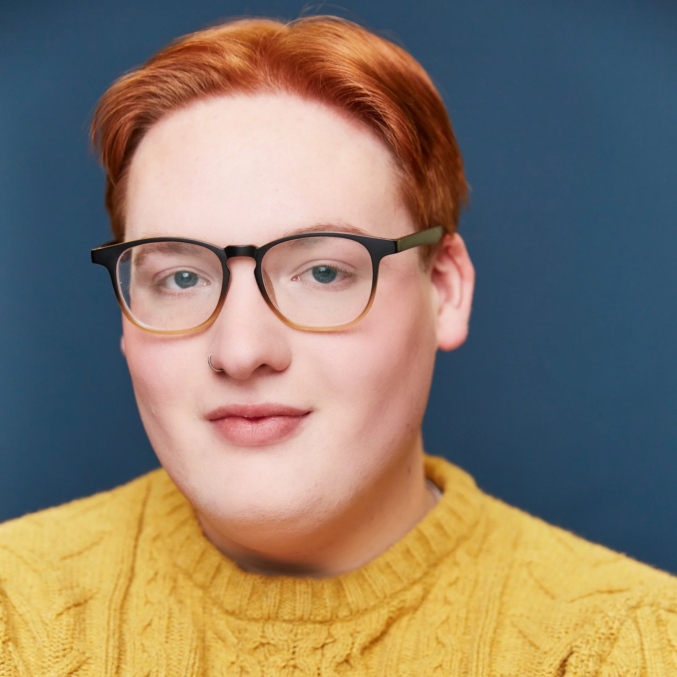 Close-up portrait of a person with short red hair, glasses, a nose ring, and wearing a yellow knit sweater against a blue background.