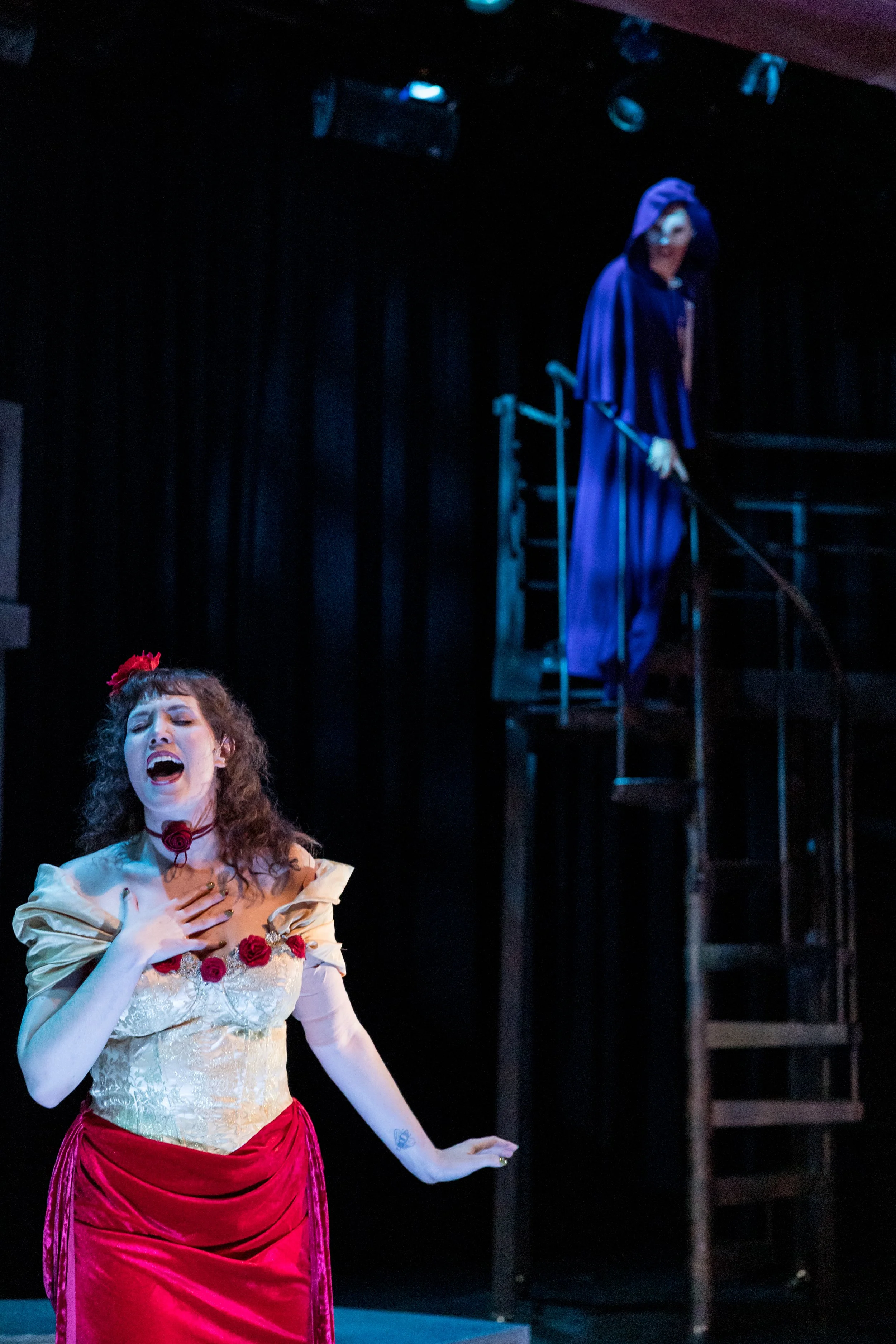 A theatrical scene with a woman singing passionately in a costume inspired by Frida Kahlo, and a man in a dark cape and hood standing on a staircase in the background.