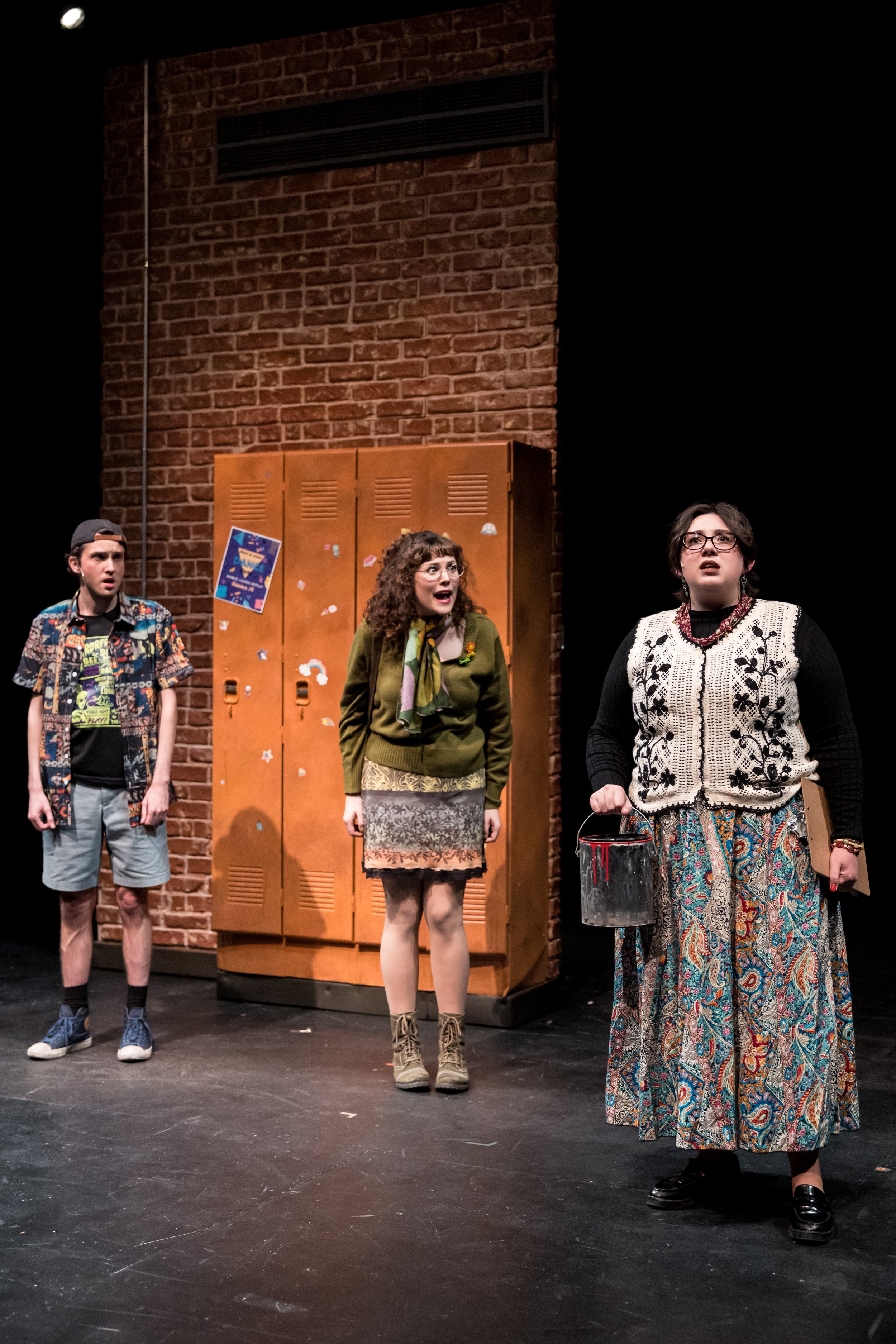 Three young people performing on stage with a brick wall and lockers in the background, appearing surprised or engaged.