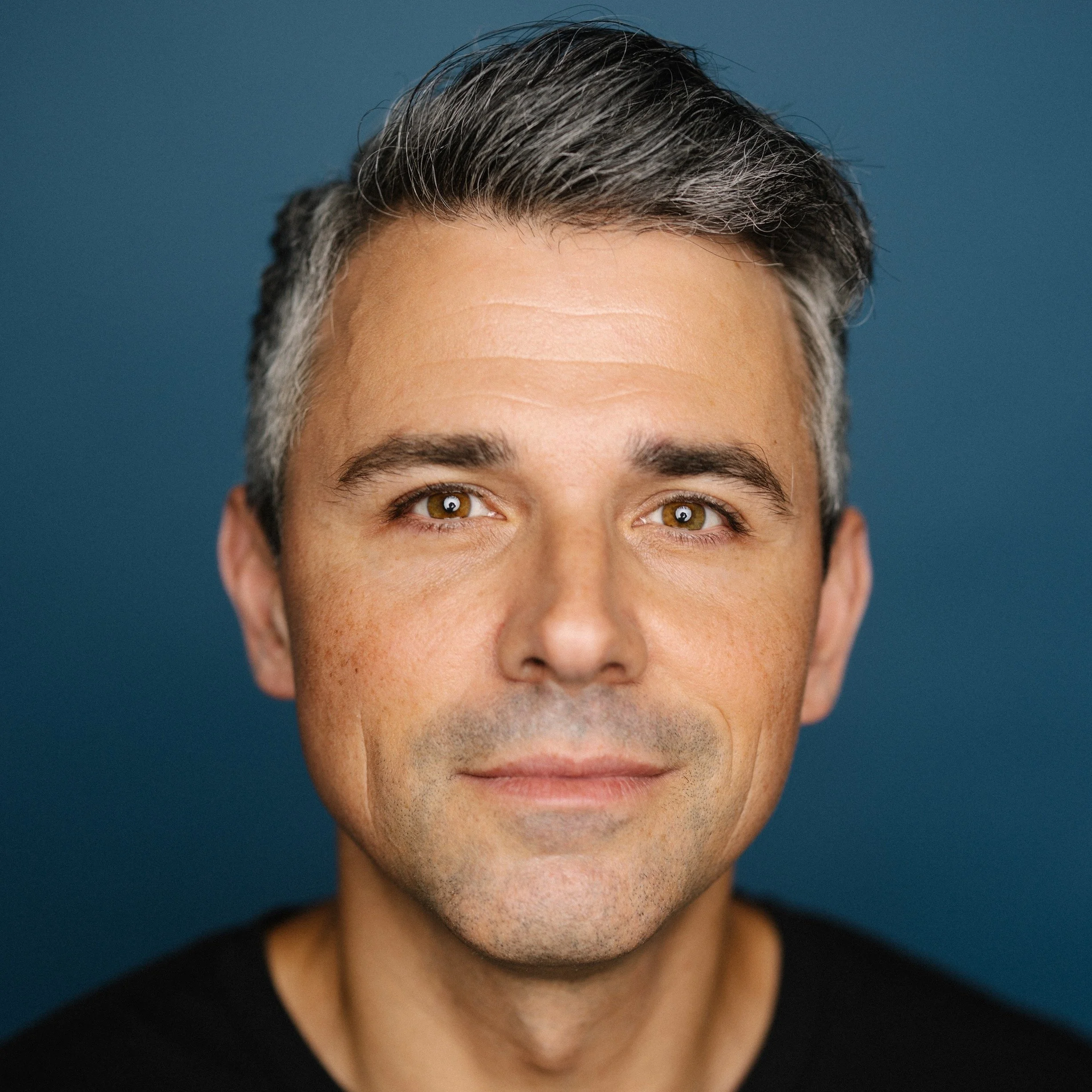 Close-up headshot of a middle-aged man with short, gray hair, hazel eyes, and light skin, smiling slightly against a solid blue background.