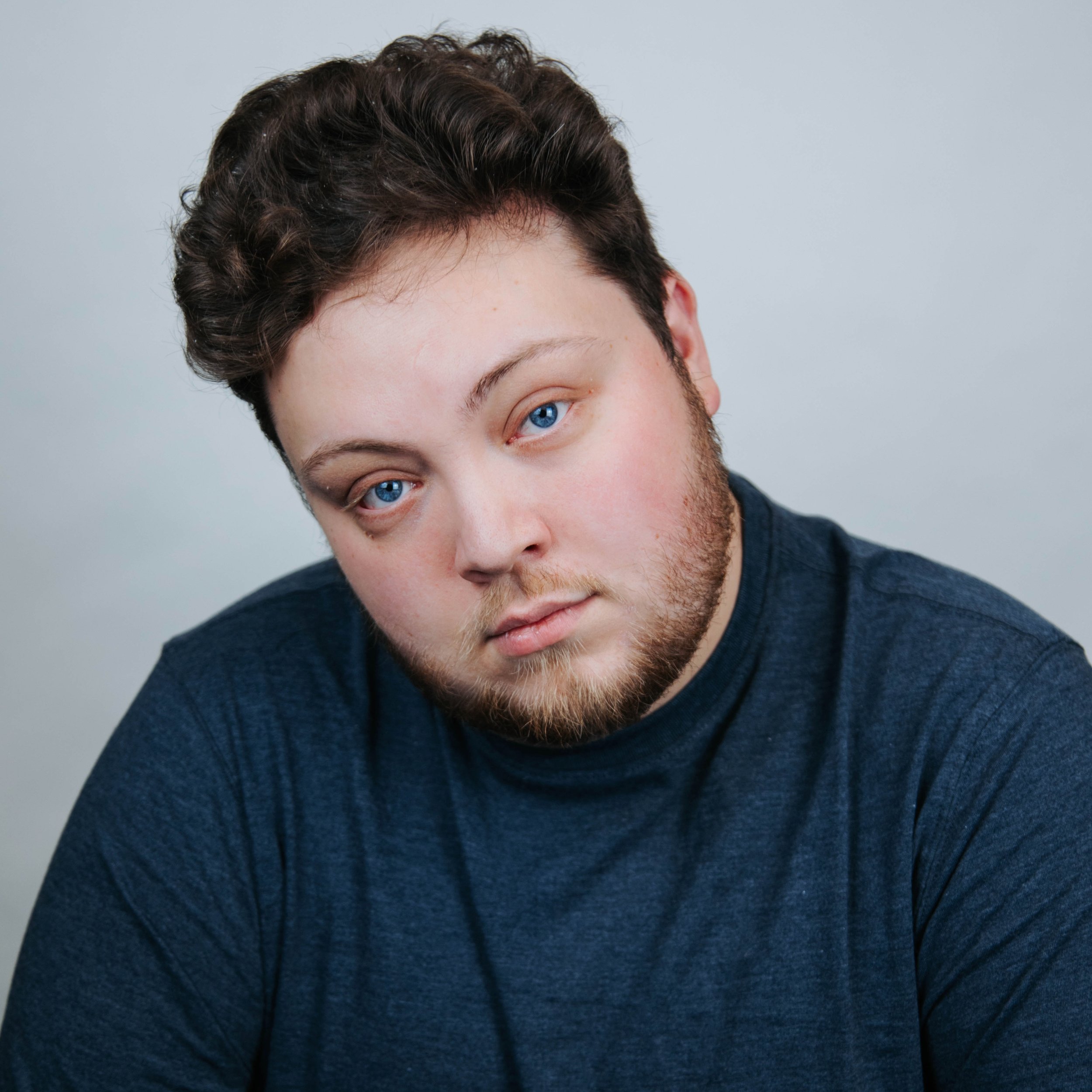 Close-up of a young man with fair skin, blue eyes, short dark brown hair, and a beard, wearing a dark blue T-shirt, looking directly at the camera with a neutral expression, against a plain light gray background.