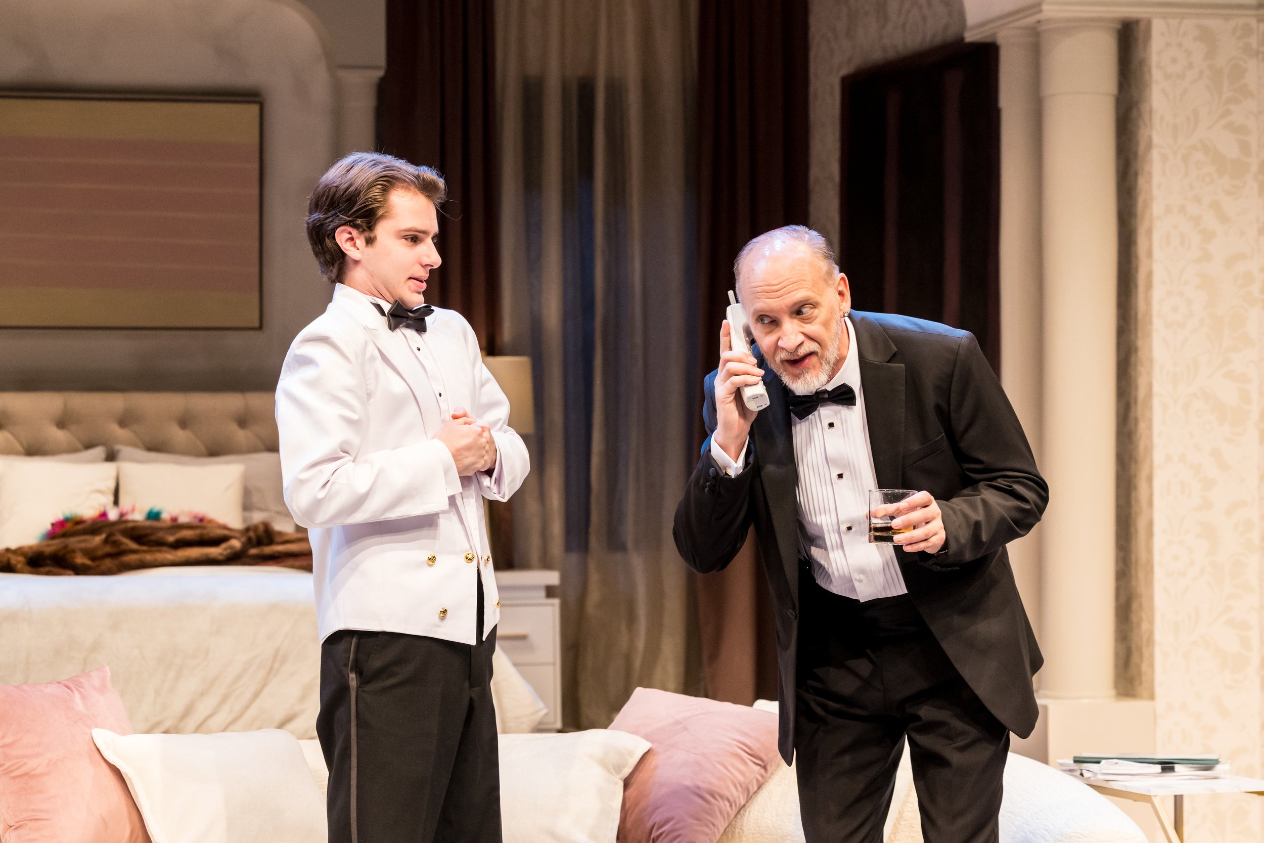 Two men dressed as waiters in tuxedos with bow ties, one older with a beard holding a phone and a drink, the other younger with a clean-shaven face, standing in a fancy room with a bed and curtains.