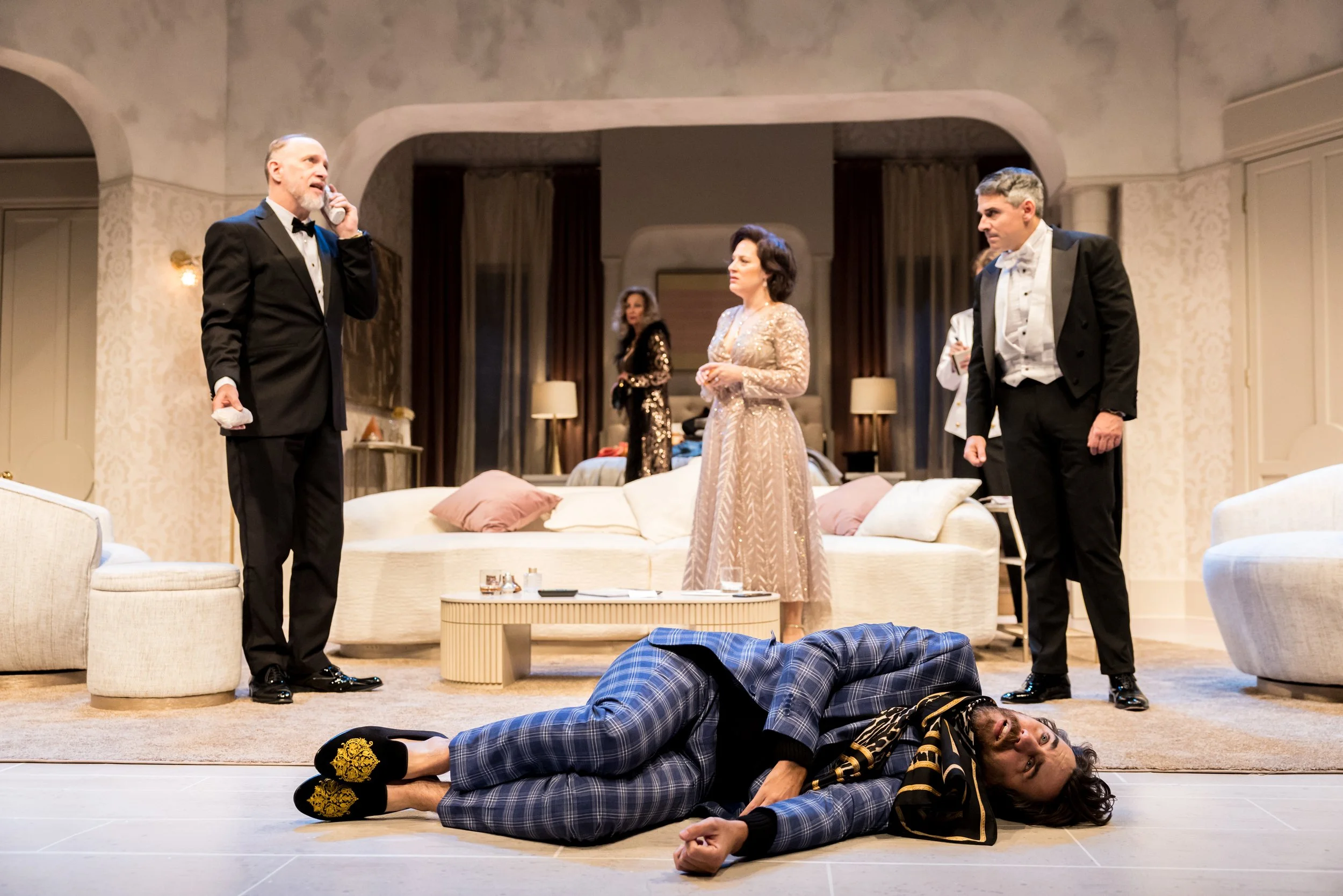 A theatrical stage scene in a luxurious living room, with five actors dressed in formal attire. One woman is lying on the floor in front, wearing plaid pajamas and slippers, appearing distressed. The other four standing are in tuxedos and a gown, wit