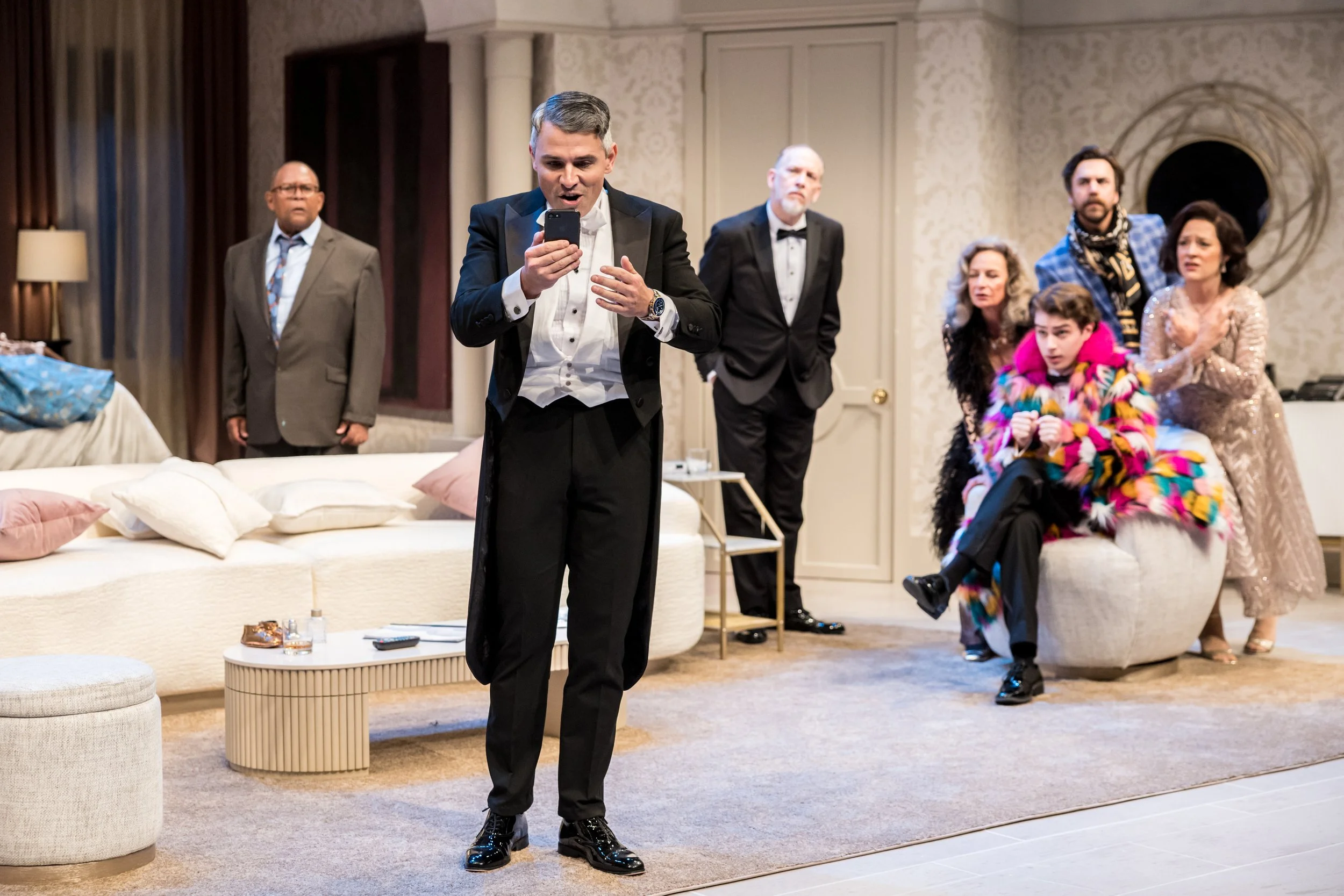Group of people in a living room, with one man in tuxedo looking at his phone and the others reacting with surprise or interest.
