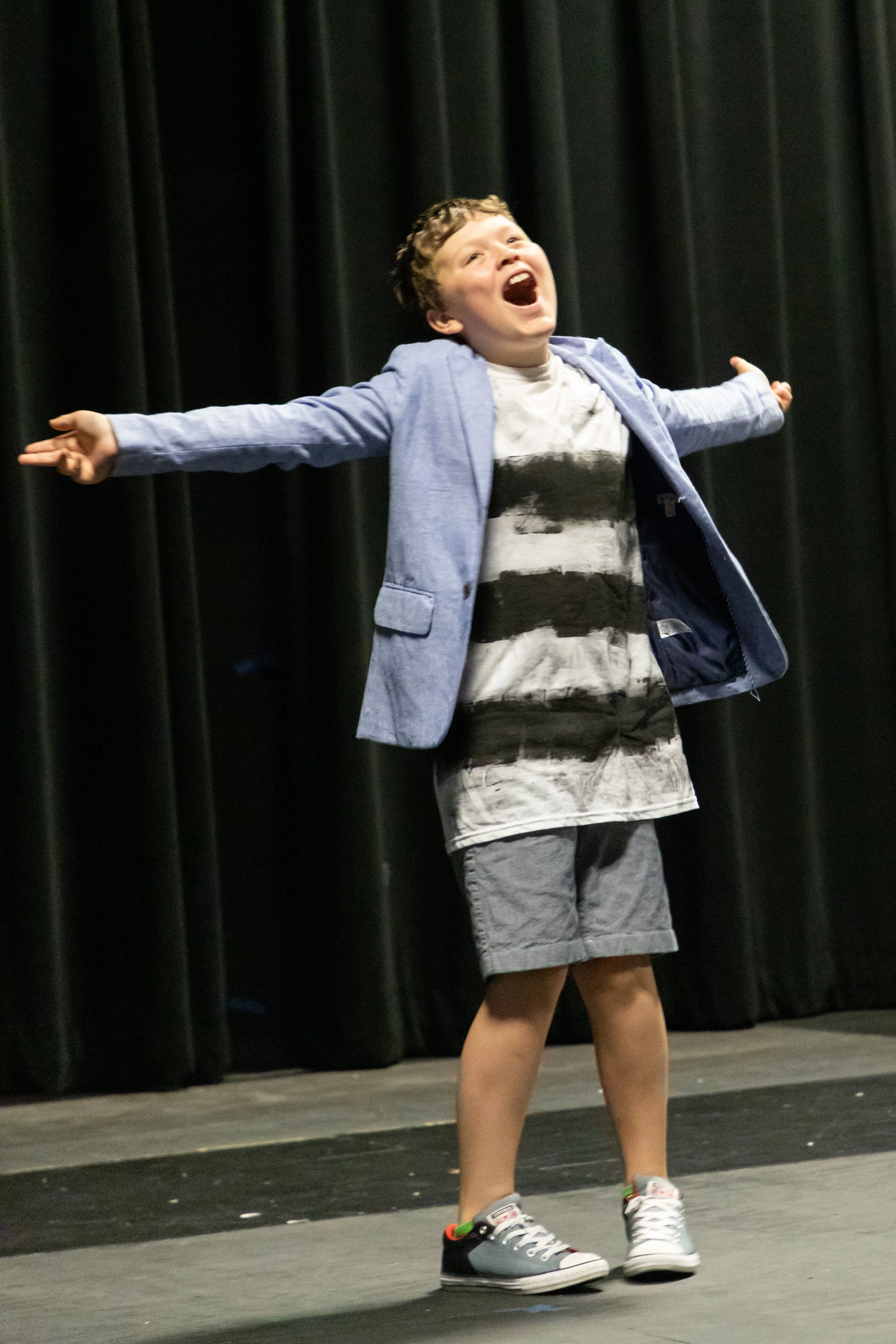 A young boy on stage with arms outstretched, wearing a blue blazer, striped shirt, shorts, and sneakers, appearing joyful and expressive against a dark curtain background.